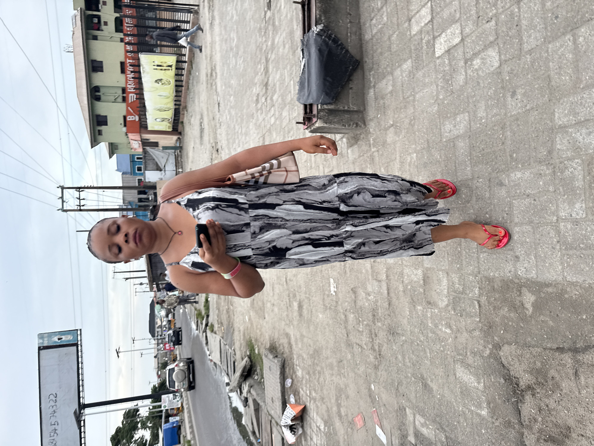 Ado, Nigeria: An individual stands on a concrete sidewalk with gray brick tiling, holding a mobile phone. They wear a gray, white, and black dress with red sandals, with a bag over their shoulder. A street runs adjacent to the sidewalk with traffic visible, and a large billboard stands to the left. Buildings are situated across the street. Power lines are visible in the background.