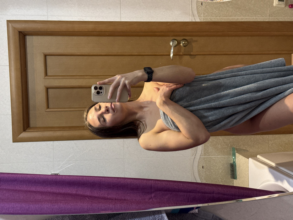 In a bathroom setting, a woman poses for a selfie in a mirror. She's wrapped in a gray towel, her upper body and arms visible. She's holding her phone in her left hand, wearing a smartwatch on her wrist. Behind her, a wooden door is visible with bathroom fixtures. A portion of a purple curtain is seen in the bottom left corner, along with a sink, hairbrush, and wall tiles. Sunlight streams into the bathroom, creating highlights on the surfaces.