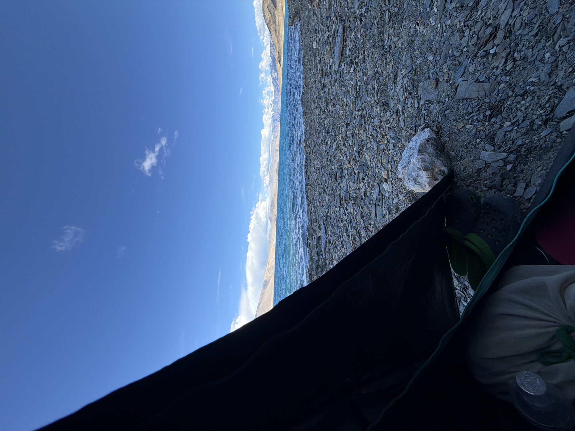 An eye-level, wide-angle, outdoor, natural light shot showcases a campsite near the serene Pangong Tso lake in Karzok, India. The foreground is dominated by the partially open flap of a dark tent, revealing a glimpse of the interior where a light-colored sleeping bag and green Crocs are visible. The ground is covered in a dense layer of grey and brown pebbles and rocks.
Beyond the campsite, the turquoise waters of Pangong Tso stretch out, rippling gently under a vast, clear blue sky dotted with a few wispy white clouds. In the distance, a majestic mountain range with snow-capped peaks forms a dramatic backdrop to the lake. The overall impression is one of tranquil isolation in a breathtaking natural setting.