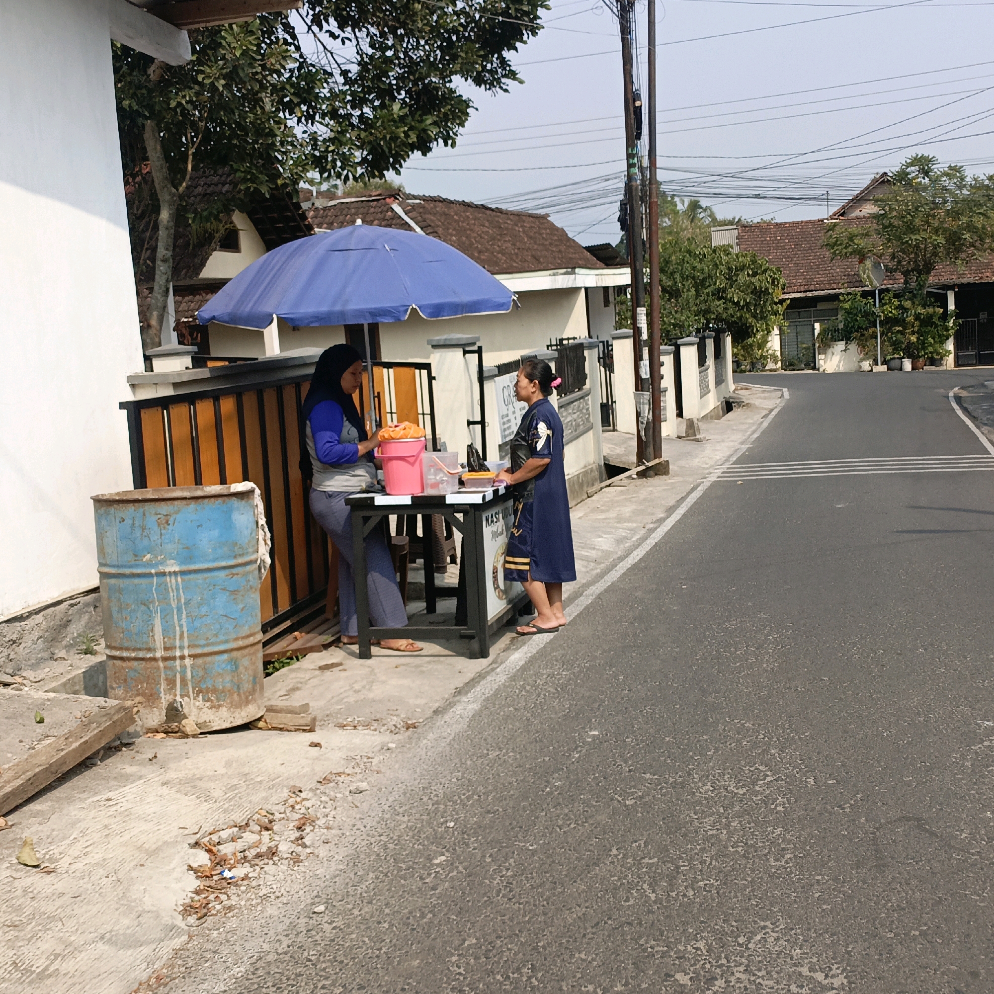 The image shows two women standing at a roadside stall in Salatiga, Indonesia. One woman, wearing a blue hijab and a blue and grey long-sleeved shirt with grey pants, is on the left. She appears to be working at the stall, with a pink bucket containing some orange items in front of her, and is looking down at the items on the table. The other woman, on the right, is wearing a dark blue dress and appears to be a customer. She is facing the stall, looking at the items and talking to the vendor. The stall itself is small and has a black and white striped sign. A blue umbrella is set up to shade the stall. A large, rusty blue barrel is positioned to the left of the stall. The scene is set on a paved roadside, with a sidewalk area in front of the stall and houses visible in the background. The sky is clear and the lighting suggests it is daytime.