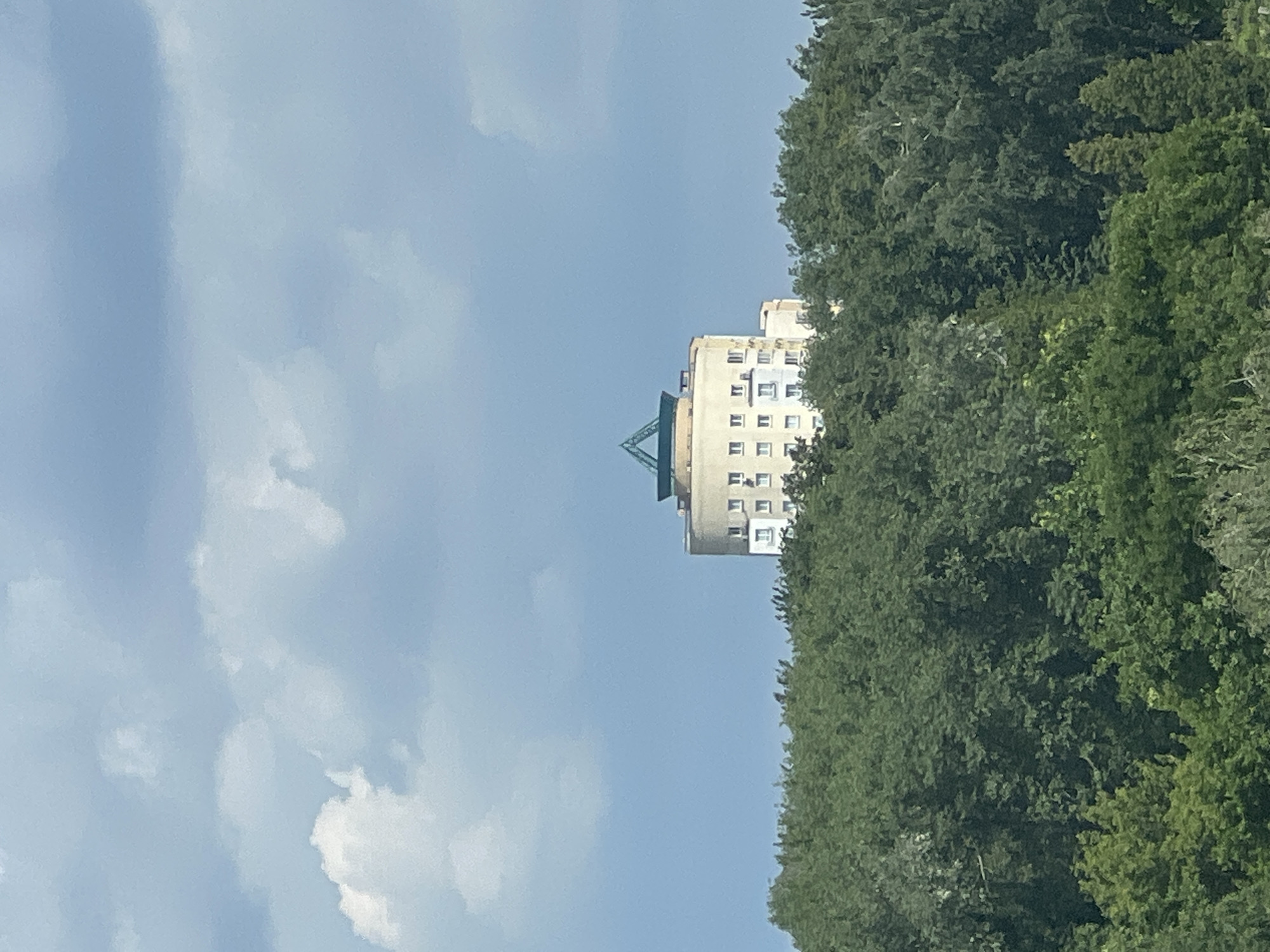 The image shows a circular apartment building with a green pyramidal roof, situated atop a dense green forest in Minsk, Belarus. The building is light-colored, with multiple windows visible. The sky above is a pale blue, dotted with fluffy white clouds. The foreground is dominated by the lush green canopy of trees.