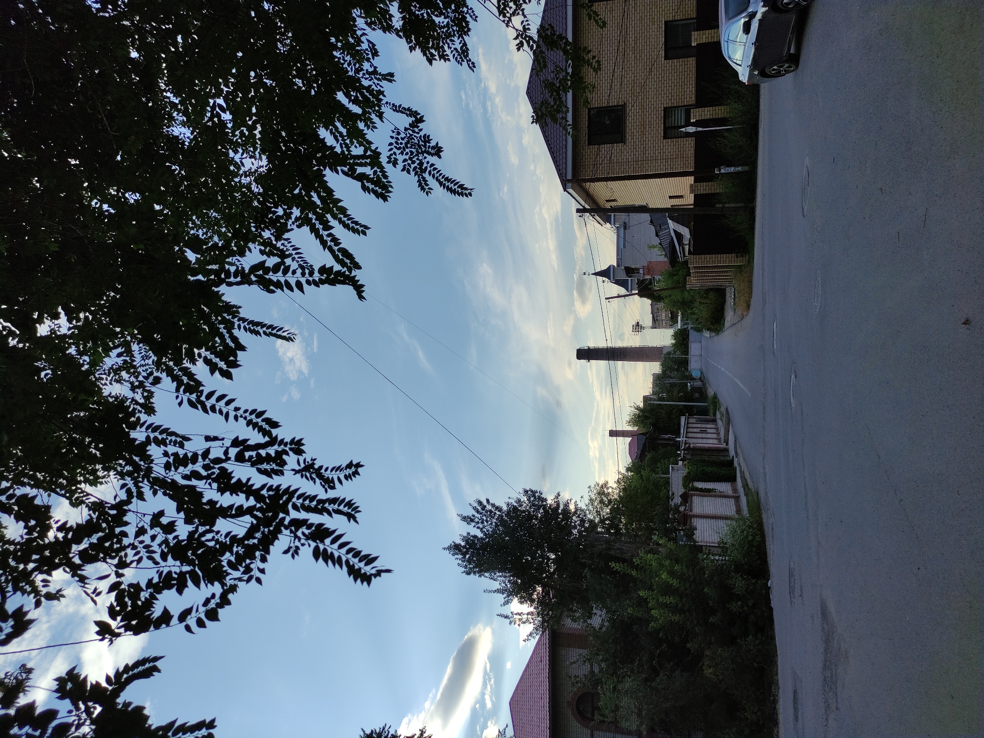 The image shows a view down a paved street in a residential area of Semey, Kazakhstan. The street is lined with fences and houses on both sides, with a prominent brick chimney visible in the distance. Trees and foliage overhang parts of the scene, and the sky is partly cloudy with a clear blue expanse. A dark-colored sedan is parked on the right side of the street. The overall scene depicts a quiet, suburban streetscape.
