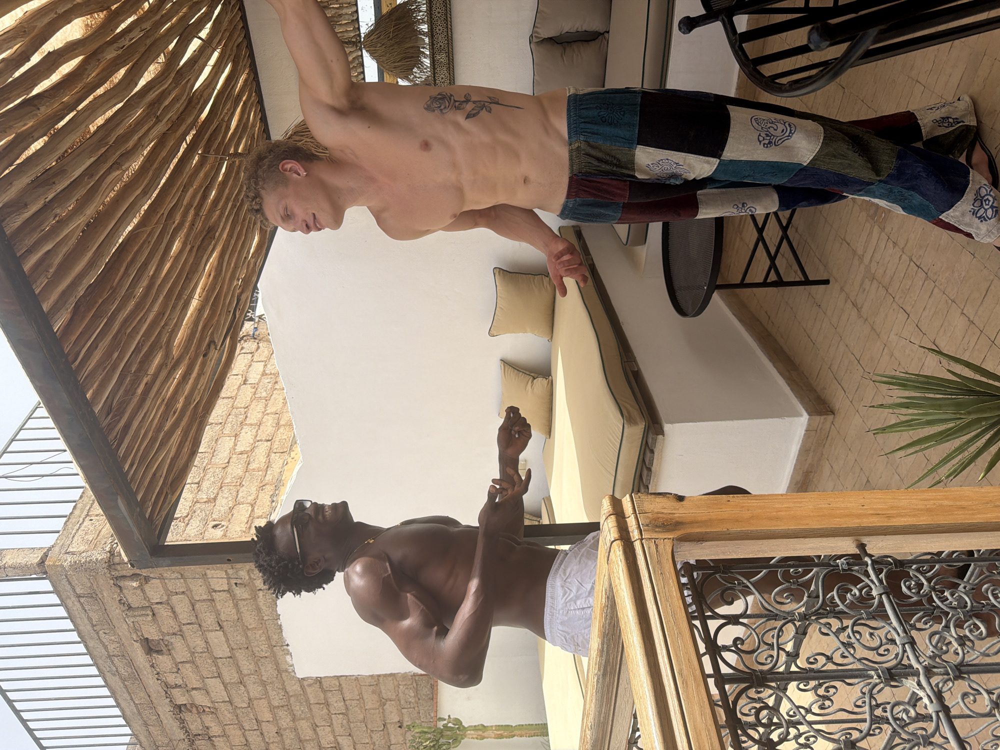 Two shirtless young men stand on an outdoor terrace in Marrakesh, Morocco. The man on the left has dark skin, short curly hair, and wears white shorts and sunglasses. He is smiling and gesturing with his hands. The man on the right has fair skin, blonde hair styled in braids, and is wearing colorful patchwork pants. He has a rose tattoo on his right rib cage. They appear to be interacting and possibly talking.
The terrace features a thatched roof overhead and brick walls. Cushioned seating areas with light-colored cushions are visible. A metal railing and decorative metalwork are present in the foreground. A potted cactus and a plant with spiky leaves are also in the scene, suggesting a warm, possibly arid environment. A small black table and chairs are visible in the background. The overall impression is a relaxed, possibly vacation setting.