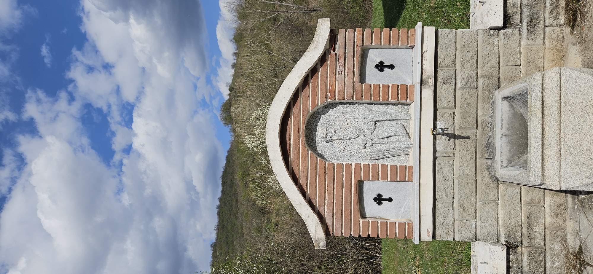 The image shows a roadside water fountain in Pastuh, Bulgaria. The fountain is constructed of red bricks, light-colored stone, and grey concrete. The structure features a curved, arched roof made of grey concrete with red bricks forming the main body of the fountain. A relief of a figure is centered beneath the arch, flanked by two stone panels with black crosses. A metal tap extends from the structure for water dispensation. To the right, a large stone basin sits atop a layered stone base, designed to catch the water. The surrounding environment includes green grass, trees with blooming white flowers, and a hillside. The sky is visible with blue patches and white clouds.