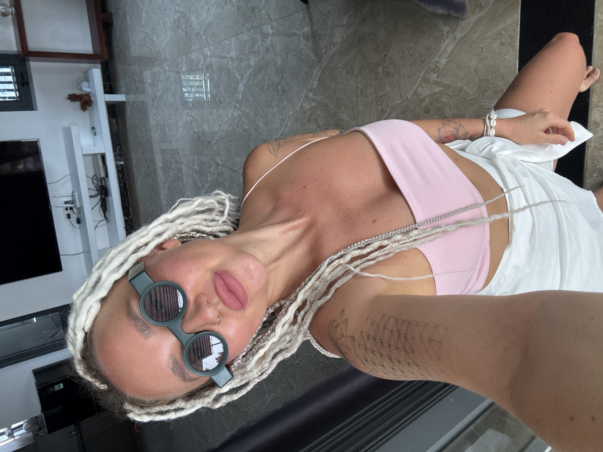 A woman with blonde dreadlocks and distinctive round, green-framed sunglasses poses for a selfie indoors. She is wearing a light pink crop top and a white wrap skirt. Tattoos are visible on her right arm and shoulder. She is kneeling on a marble-tiled floor, which reflects light and the surroundings. In the background, a television set is mounted on a white entertainment unit. The environment appears to be a modern living space, possibly an apartment in Bac Nha Trang Ward, Vietnam, given the context.