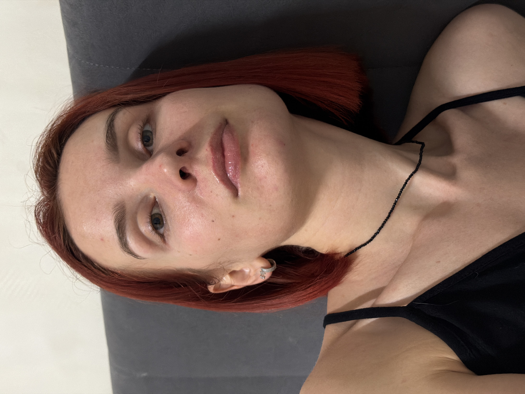 A woman with short red hair lies on a gray cushion. She wears a black tank top and a black beaded necklace. Her skin is light-toned with visible pores and minor blemishes. She has light eyebrows and gray eyes. The background consists of a white wall. There's no indication of Chaikovsky, Russia.