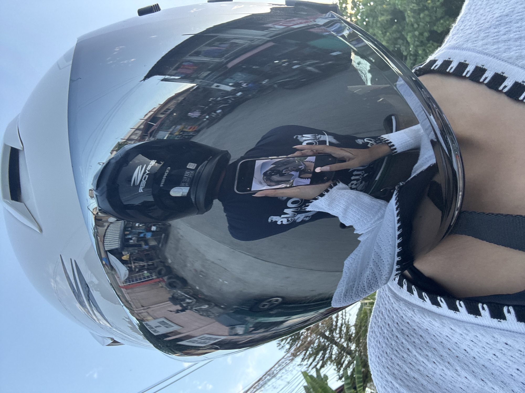 The image is a first-person perspective of a person wearing a white motorcycle helmet with a reflective visor. The visor reflects a street scene, likely in a tropical urban environment, possibly in Davao City, Philippines, due to the style of buildings and vegetation. The reflection shows a narrow street lined with buildings with open fronts, some displaying goods. There are parked motorcycles and what appear to be discarded tires. The sky is visible above the buildings. The person is holding a smartphone, taking a selfie, and their reflection is visible in the visor along with the surrounding environment. They are wearing a white knitted garment over a dark top. The overall impression is a snapshot of daily life on a street.