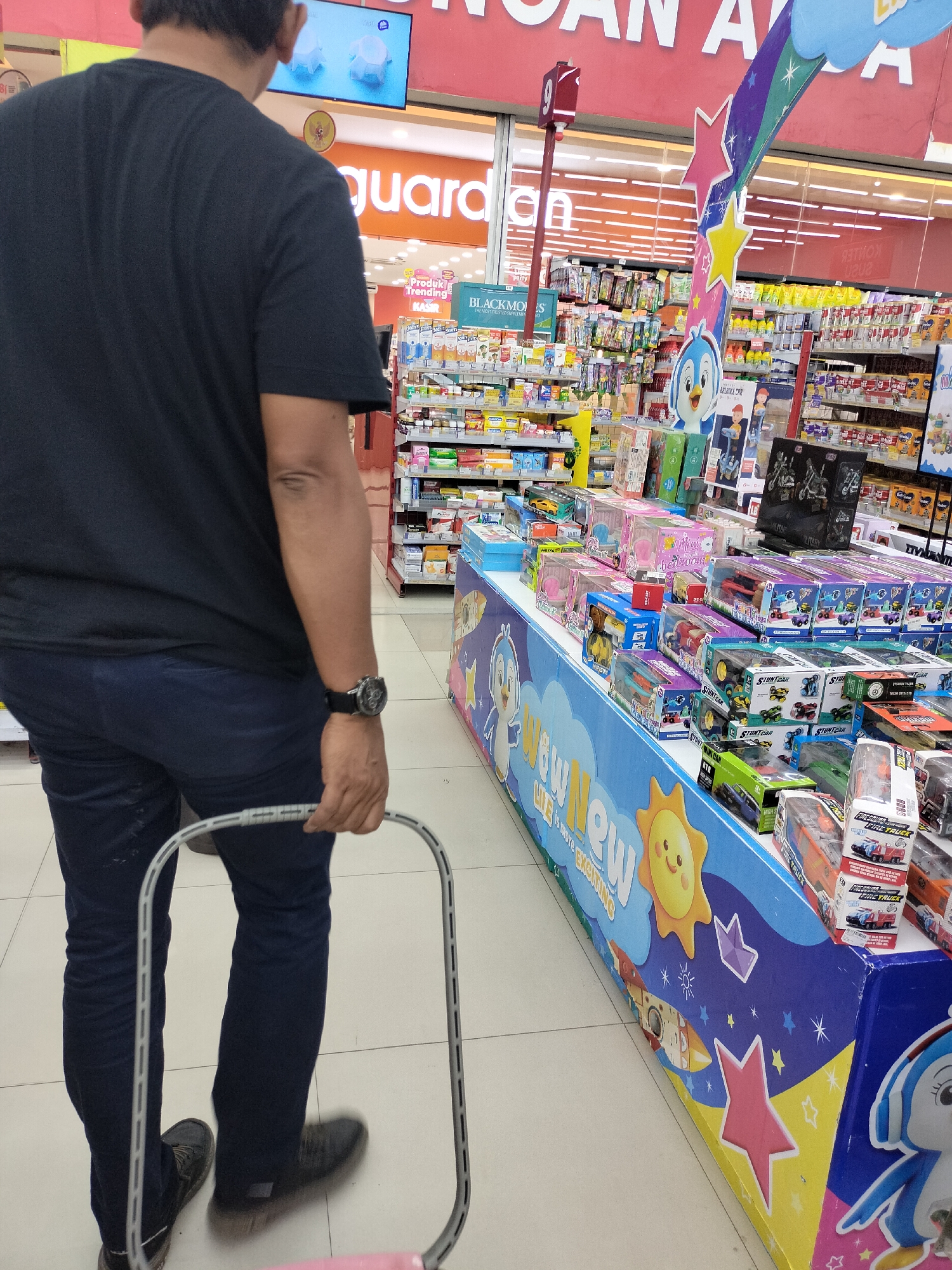 The image was taken in Langsa, Indonesia, inside a retail store, possibly a supermarket or a convenience store. The store appears well-lit and stocked with various products. In the foreground, a person is seen from the back, wearing a black t-shirt and dark blue jeans. They are holding onto the handle of a shopping basket, indicating they are in the process of shopping. The person is wearing a watch on their left wrist.
The background of the image reveals rows of shelves filled with packaged goods. The shelves display a variety of items, including what appear to be health and beauty products, snacks, and toys. There's a prominent display of toy cars and other boxed toys on a counter in the middle ground, decorated with cartoon characters and colorful graphics. A large sign with red lettering that says "GUAN A" is visible above the shelves, likely part of the store's name or a promotional banner. A digital screen displaying images of what seem to be household items is mounted high on the wall. The floor is tiled with a light-colored pattern. The overall impression is of a typical retail environment.