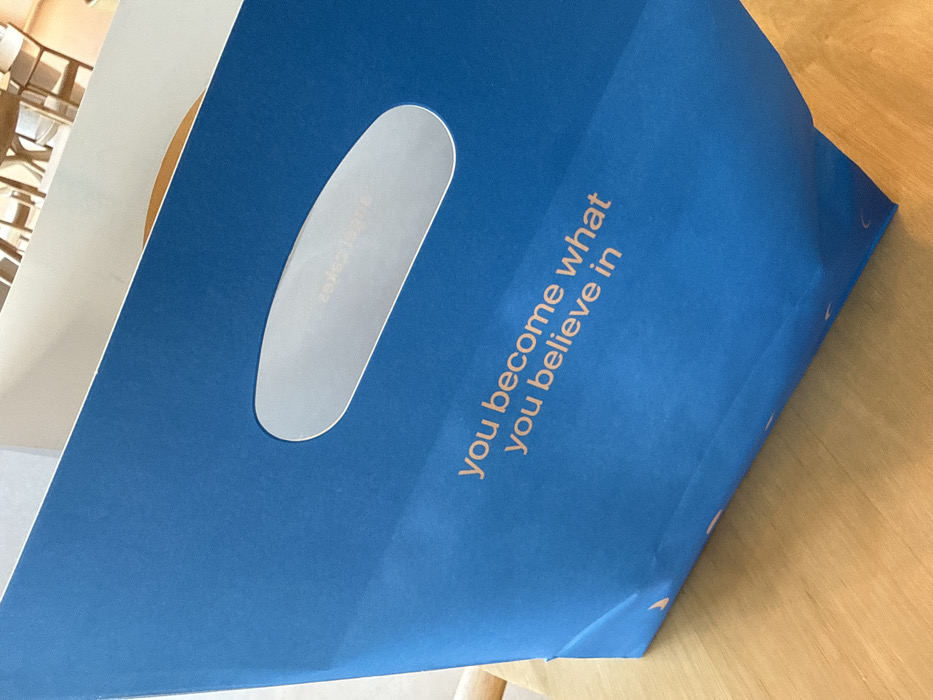 A royal blue paper shopping bag, bearing the slogan "you become what you believe in" in light orange text, rests on a light-colored wooden table surface. The bag features an oval-shaped die-cut handle, within which a faint, mirrored reflection of "Angelic Cakes" is visible. The background, out of focus, reveals an indoor setting with light-toned wooden chairs and tables, suggesting a cafe or bakery environment. The item originates from Angelic Cakes, located in the city of Dubai, United Arab Emirates. No individuals or specific actions are depicted.