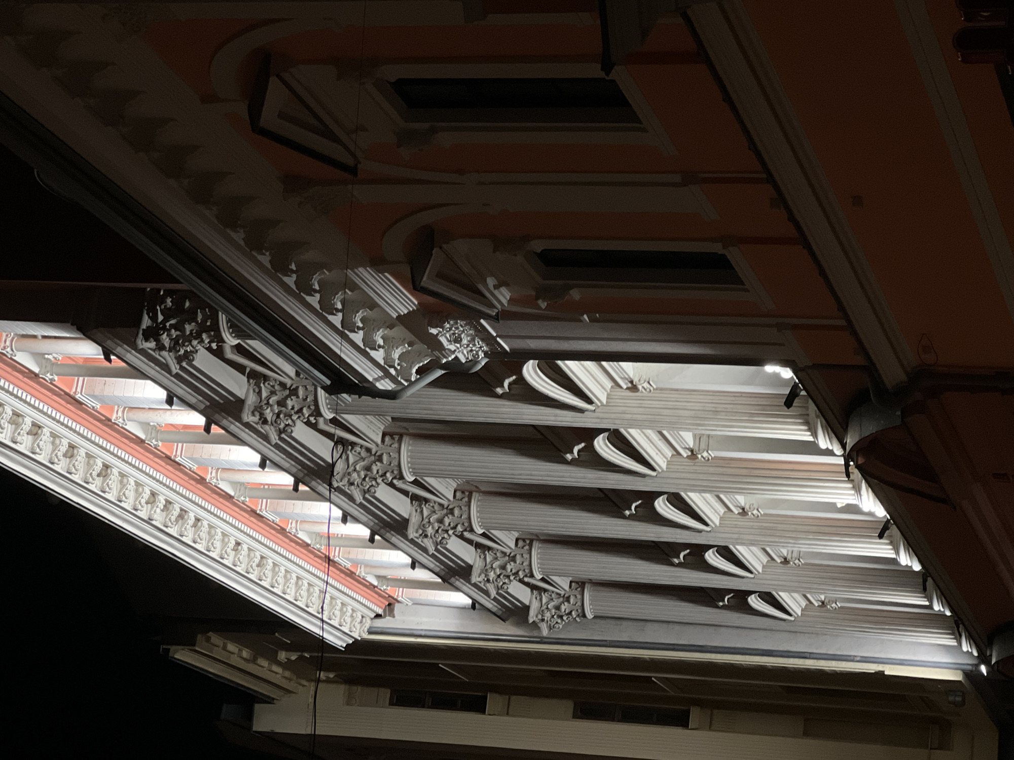 The image shows the facade of a historic building at night, illuminated by artificial lighting. The building features classical architectural elements, including Corinthian columns with ornate capitals, a decorative frieze, and arched window openings. The facade is painted in shades of orange and white. The perspective is angled, looking up at the building from below. There are no people visible in the image. The lighting creates strong shadows and highlights, emphasizing the architectural details. The surrounding environment appears to be a city street, with darkness visible in the background, suggesting it is nighttime. Based on the architectural style and typical lighting of such buildings, this is likely the facade of the Bulgarian National Bank building in Plovdiv, Bulgaria, which is known for its classical architecture and prominent location.