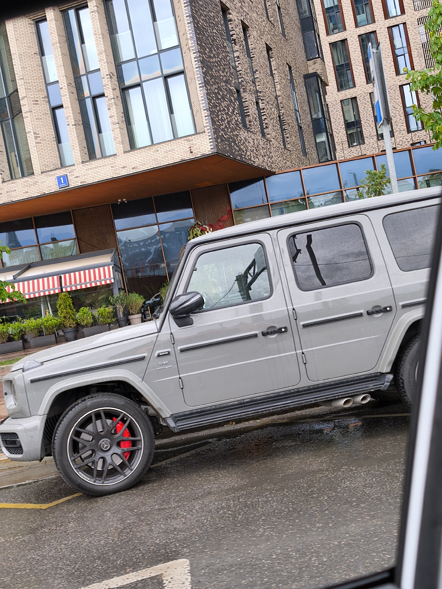 The image shows a gray Mercedes-Benz G-Class parked on a wet street in Moscow, Russia. The vehicle is positioned in front of a modern building with a restaurant awning visible behind it. The building's architecture includes a combination of brick and glass. The street is wet, indicating recent rainfall. There are no people visible in the immediate vicinity of the car. The focus is on the vehicle and its surroundings, emphasizing the urban environment.