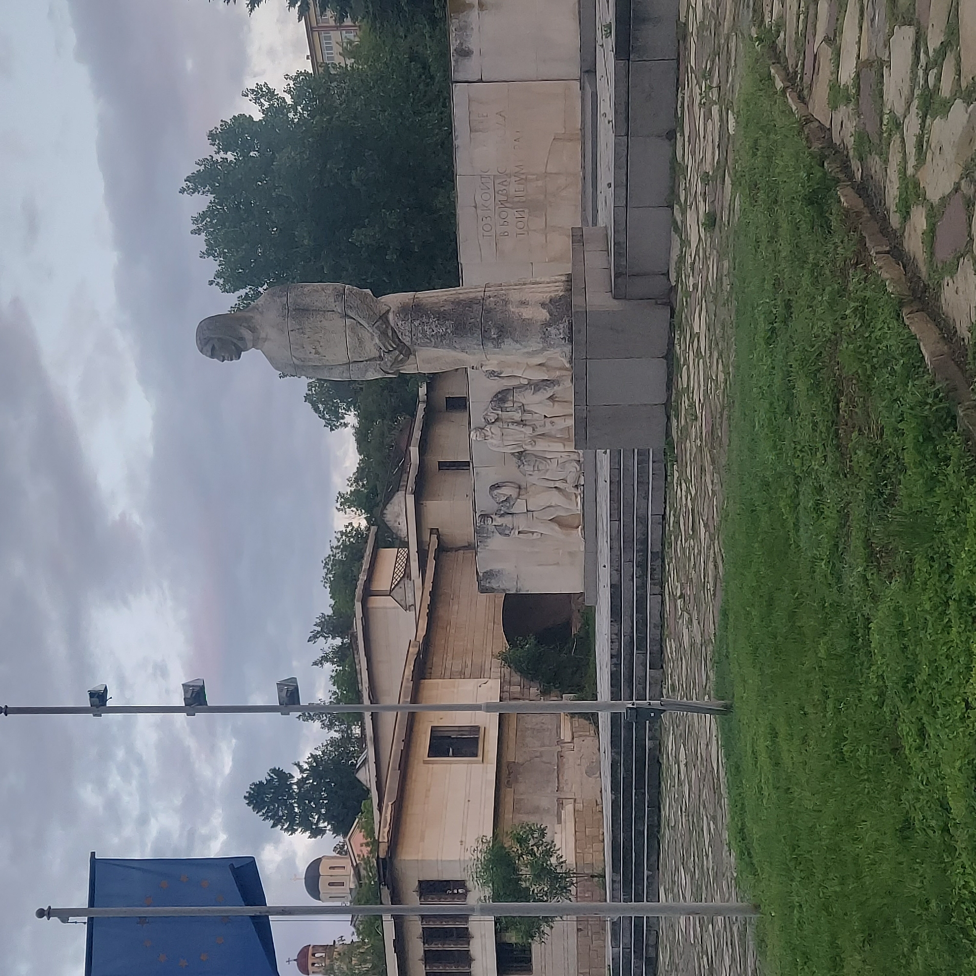 The image depicts a monument, potentially a war memorial, in Strazhitsa, Bulgaria. The monument consists of a stone statue of a draped figure, likely a woman, positioned above a relief depicting a group of individuals. The monument is constructed from light-colored stone. The statue is situated on a stepped platform with an inscription in Cyrillic on the side.
The setting includes green grass and paving stones surrounding the memorial. Buildings are visible behind the monument, including what appears to be a church or other historical structure with domes. Utility poles and the European Union flag are visible in the foreground. The sky is overcast.