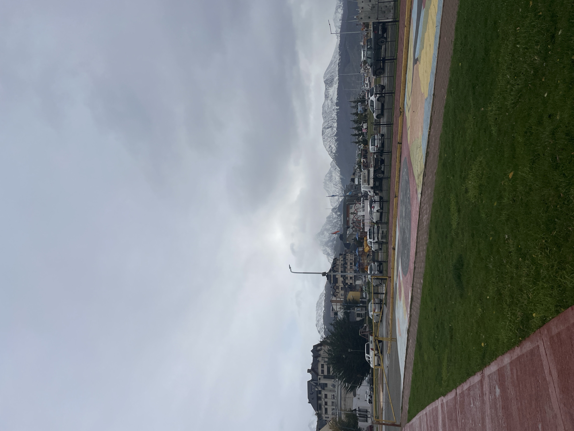 The image shows a landscape in Ushuaia, Argentina, under a cloudy sky. In the background are mountains with visible snow patches. In the midground is a town with buildings of varying heights and colors, interspersed with trees. A street runs along the middle ground, populated with cars. In the foreground, there is a red-painted curb, and a grassy area, which is directly next to a ground mural. The image appears to have been taken in a public space overlooking the town. There are no visible people in the image.