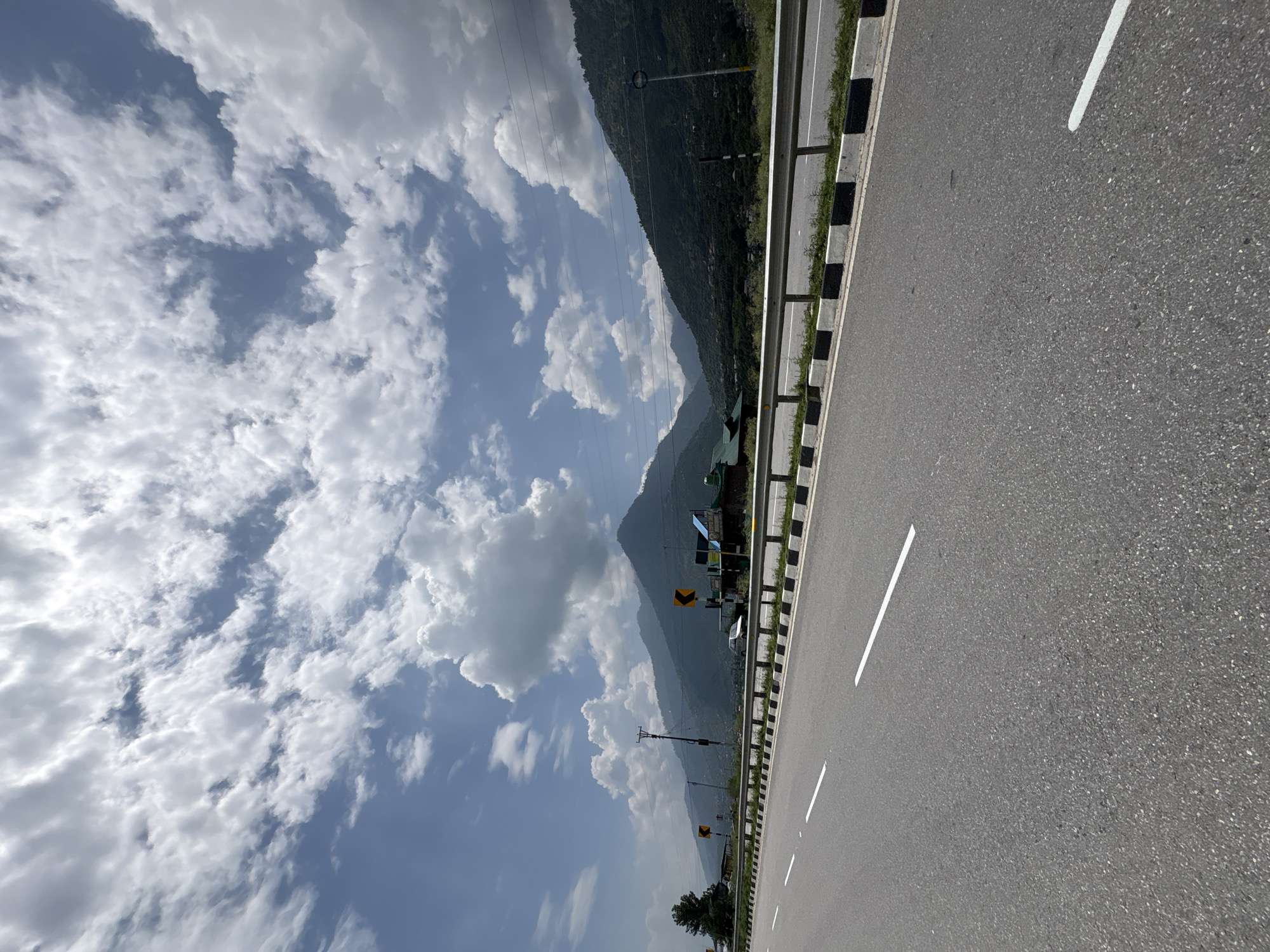 The image captures a daytime view of a highway with mountains in the background, likely in the vicinity of Pirdi, India. The highway is paved with asphalt and features white lane markings and a black and white striped barrier along the side. Green vegetation borders the highway, and several mountainous hills covered in trees rise in the distance. The sky is partly cloudy with fluffy white cumulus clouds against a blue backdrop. Signs and utility poles are visible along the roadside. The overall scene depicts a scenic mountain highway under a partly cloudy sky.