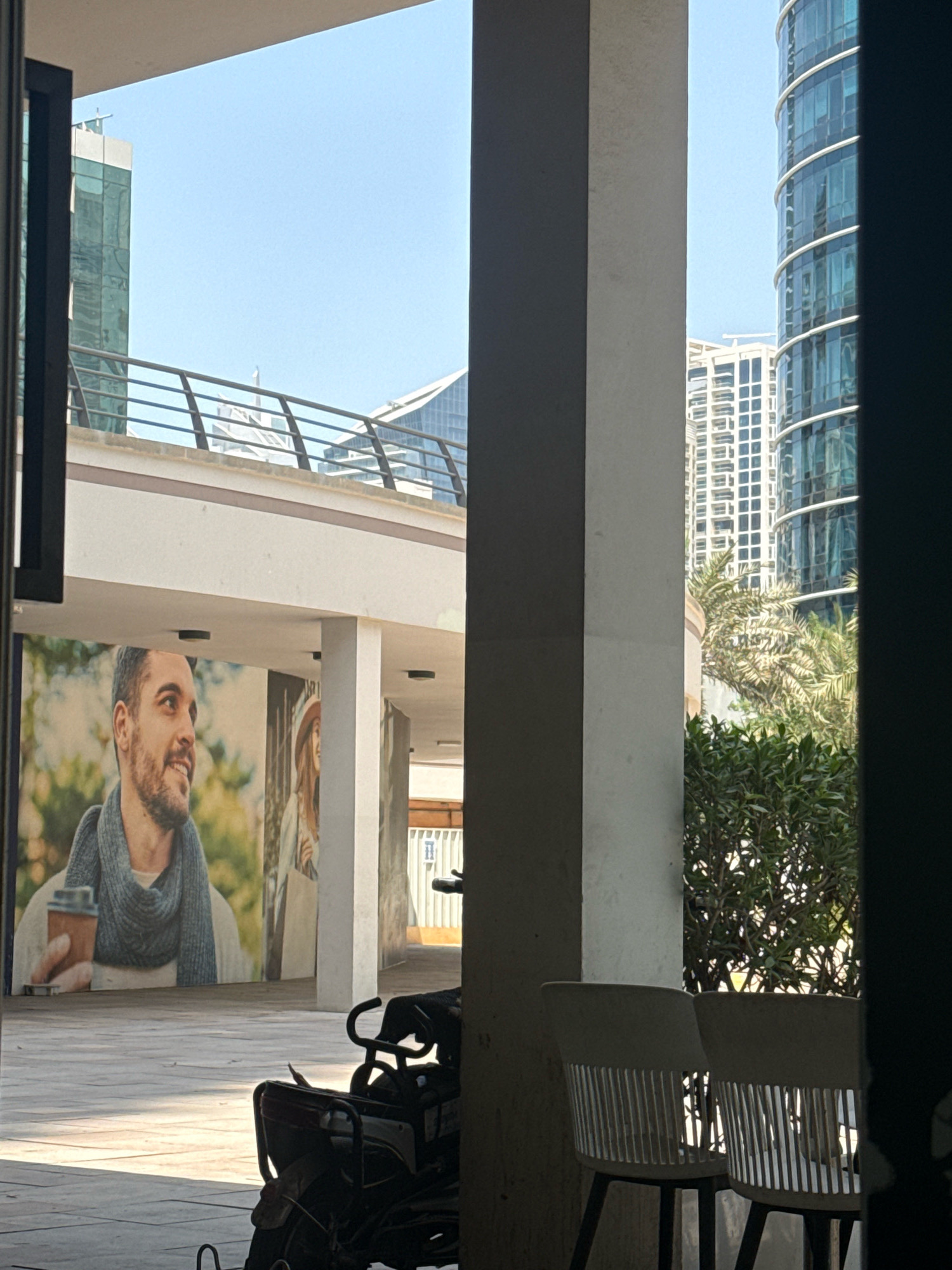 The image shows an outdoor seating area in Dubai, United Arab Emirates, with a view of modern skyscrapers. In the foreground, two chairs with slatted backs and dark legs are positioned on a paved ground. To the left, a mural of a man with a beard, wearing a scarf and holding a coffee cup, is visible on a wall. Behind the chairs and to the right, lush green bushes and palm trees are present. In the background, several tall buildings with glass facades, characteristic of Dubai's architecture, dominate the skyline under a clear blue sky. A motorcycle is parked in the middle ground. The scene suggests a commercial or public space, possibly a cafe or a plaza, designed to offer both seating and a scenic urban backdrop.