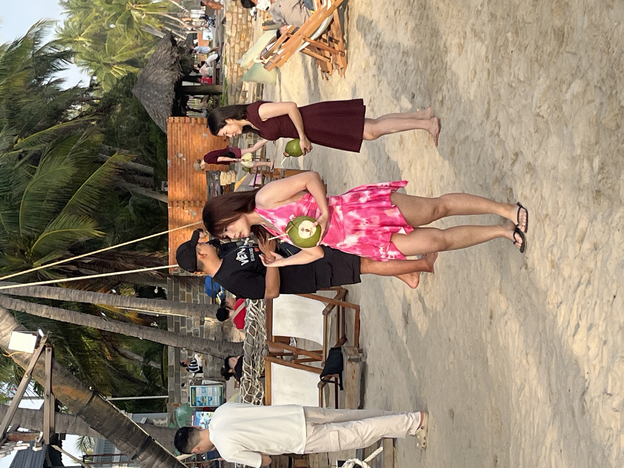 The image depicts a beach scene in Mui Ne, Vietnam. In the foreground, a woman in a pink, tie-dye dress holds a coconut with a straw. To her right, another woman in a dark red dress also holds a coconut. Behind them, a man in a black t-shirt and shorts stands facing away from the camera. Further back, a man in a white t-shirt and beige pants is visible. The background shows palm trees, a thatched-roof hut, and people relaxing on beach chairs. The environment is sandy, with evidence of typical beach amenities and vegetation.