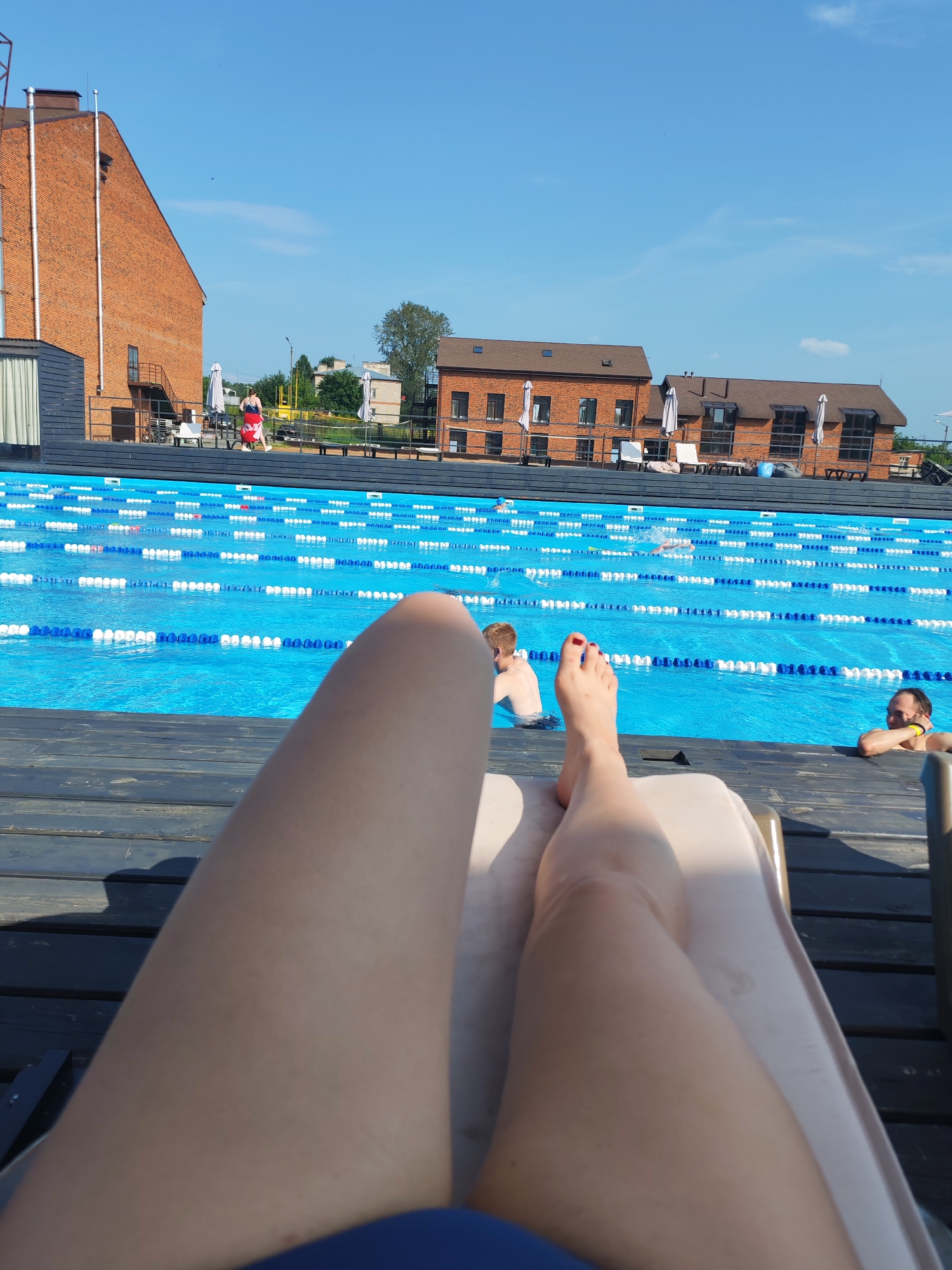 The image depicts a sunny day at an outdoor swimming pool complex, likely located in Pirogovo, Russia. The foreground is dominated by the legs of a person relaxing on a lounge chair, with their feet prominently displayed. The pool itself is a vibrant blue, divided into lanes by white and blue buoys, suggesting it is set up for swimming laps or training.
Several people are visible in and around the pool. In the middle ground, a person is swimming in one of the lanes. To the left, a person with red shorts is walking on the deck, and further back, a person with blonde hair is in the water. On the right side of the pool, a man is leaning on the edge, looking towards the camera.
The background shows several brick buildings with arched windows and dark roofs, characteristic of modern architecture. White umbrellas and lounge chairs are scattered around the poolside, indicating a leisure and recreational area. The sky is clear and blue with a few wispy clouds, and the bright sunlight suggests it is daytime, likely late morning or early afternoon. The overall atmosphere appears relaxed and summery. No visible text is present in the image.