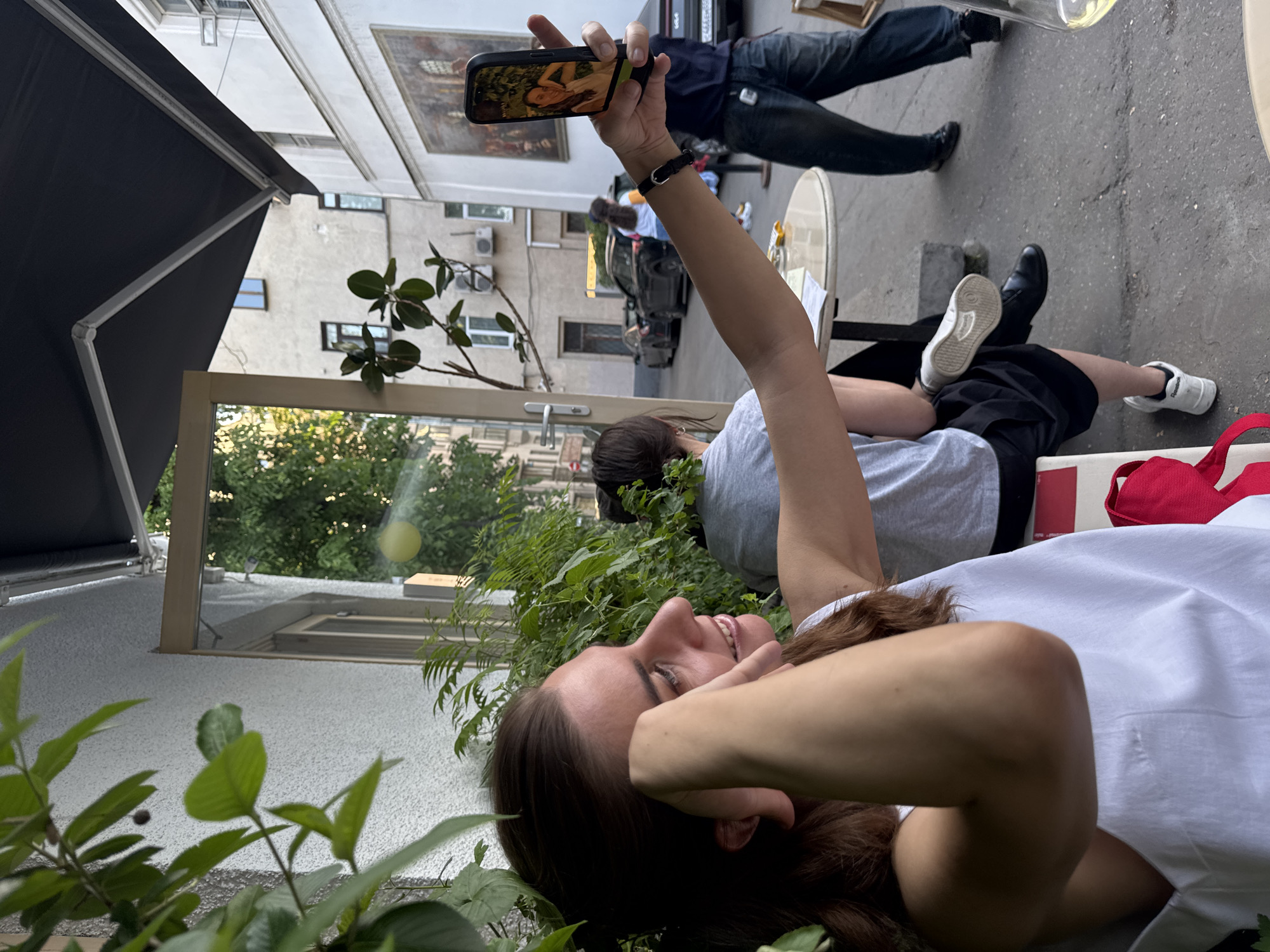 A woman is lying down in a grassy area, possibly a courtyard. She is holding a smartphone up in the air, apparently taking a photo or recording video. On the screen of the phone, a woman is depicted, likely from artwork. Other people are present in the background, including a person lying down and a standing person wearing jeans. Tables and chairs are visible, suggesting an outdoor seating area of a restaurant or cafe. The scene takes place in Moscow, Russia. The architecture of the surrounding buildings matches Moscow’s urban style.