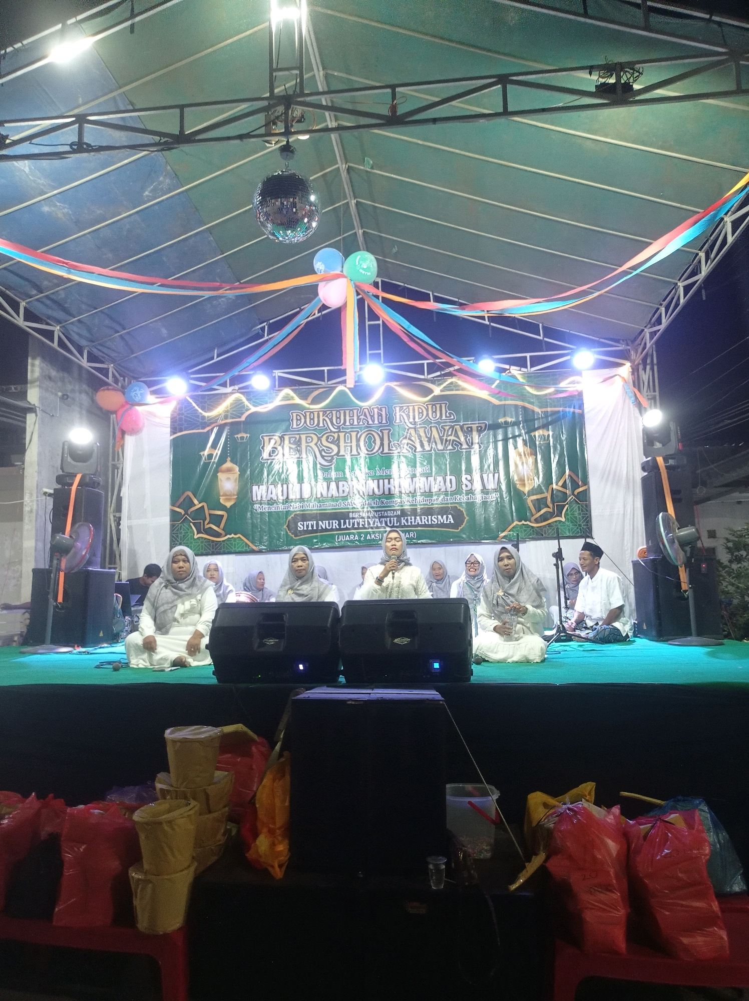 The image depicts an outdoor stage event at night, likely in the city of Tuban, Indonesia. A green banner with text in Indonesian, promoting a religious gathering or celebration, hangs prominently on the stage. Several people are seated on the stage, dressed in white religious attire, with some appearing to be speaking or leading the event. A man in the center is speaking into a microphone. Decorative elements like colorful streamers and balloons are visible. In the foreground, several bags and containers are placed on red plastic tables. Large speakers are positioned on the stage and around the venue. A disco ball is suspended from the stage's roof. The overall ambiance suggests a community event or religious ceremony.
