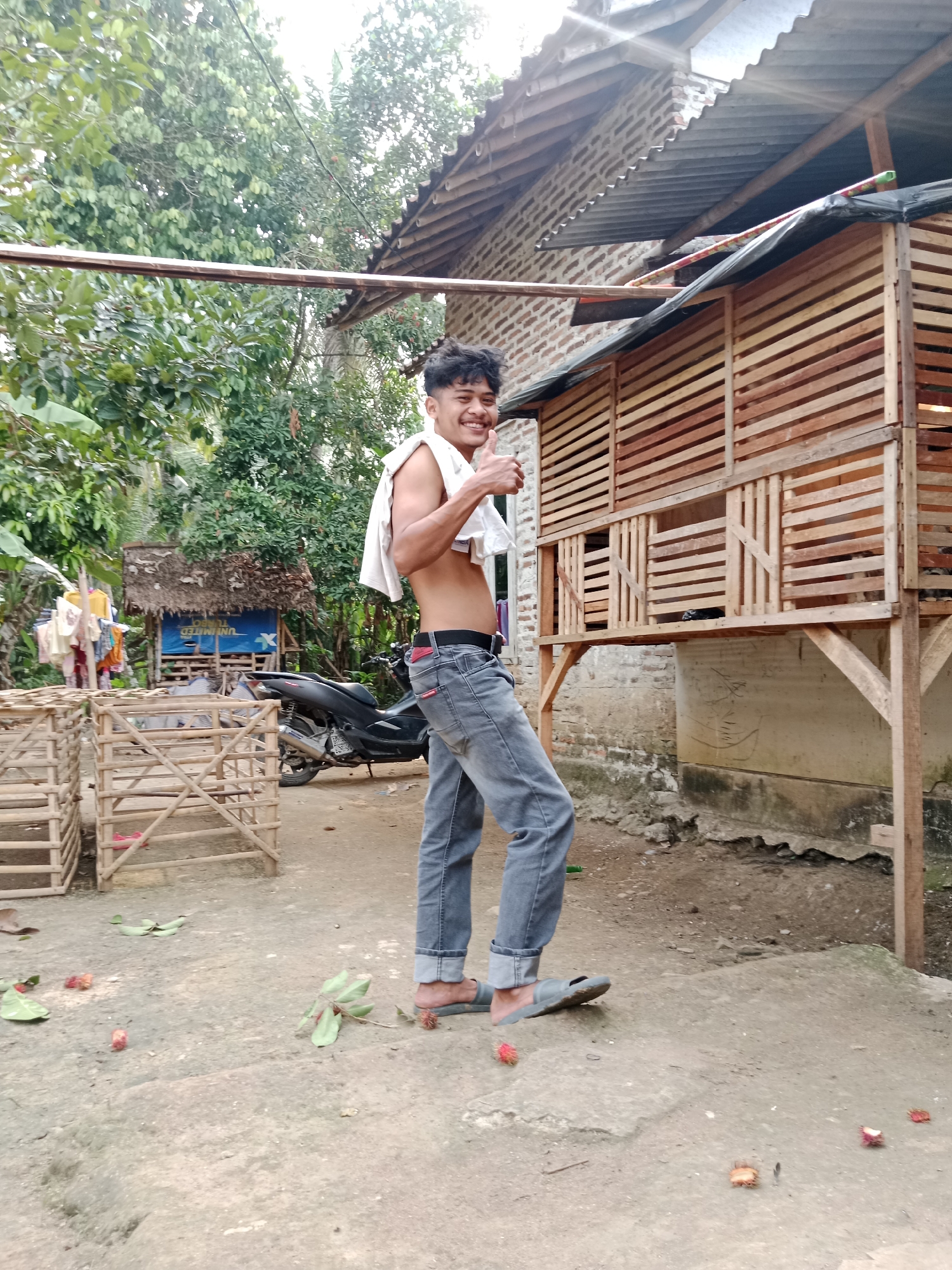 In Parage, Indonesia, a young man stands outdoors, giving a thumbs-up. He is shirtless, wearing a white towel draped over his shoulders, and grey jeans. The background features a wooden structure, a motorcycle, and lush greenery. Scattered on the ground are rambutan fruits and leaves. The scene suggests a rural or semi-rural environment, possibly a farm or residential area.