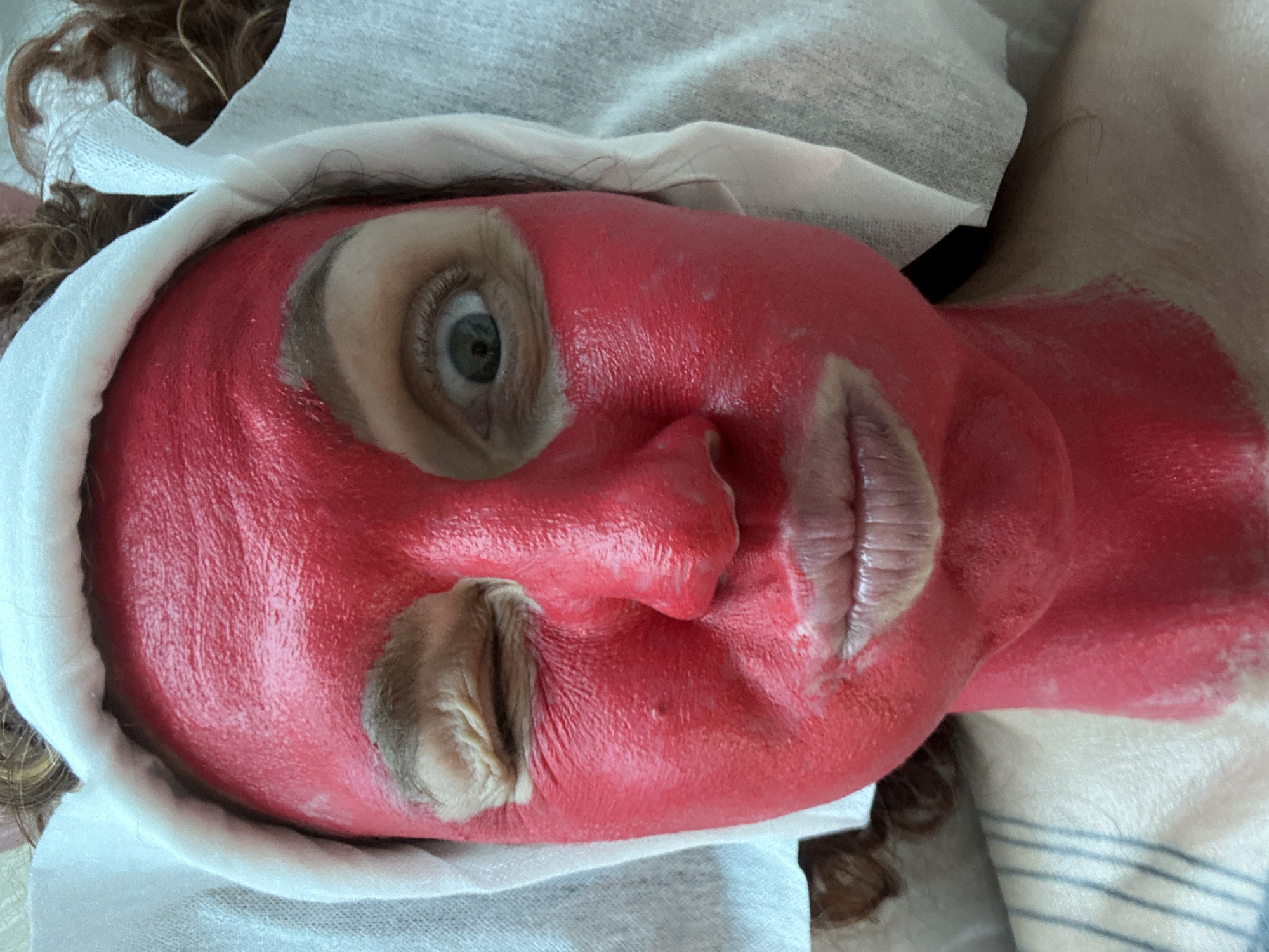 The image shows a person lying on a surface covered with a white and blue striped cloth. The person's face is covered in a thick, bright red substance, with areas around the eye and mouth uncovered. A white cloth is draped around the person's head. No details in the image connect it to the city of Sochi, Russia.