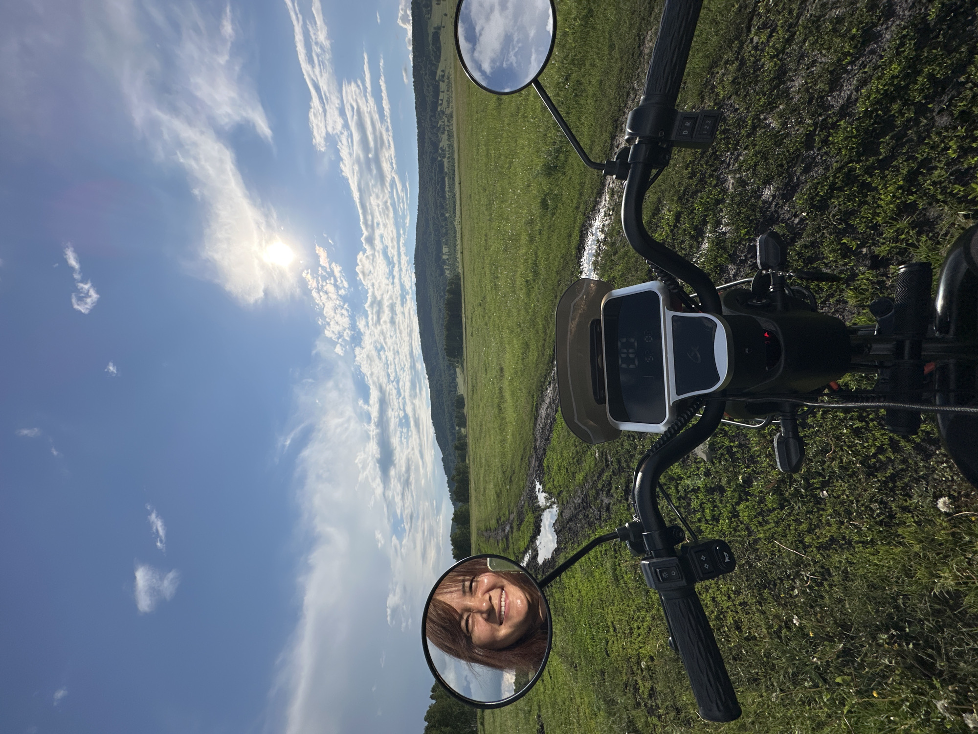 A first-person perspective shot captures a woman's reflection in a side mirror of an electric scooter. She is smiling and looking directly at the camera. The scooter is positioned on a muddy path in a lush green field. In the background, a tree line and rolling hills form the horizon, under a bright, sunny sky with scattered clouds. The overall scene suggests an outdoor adventure in a rural or natural setting.