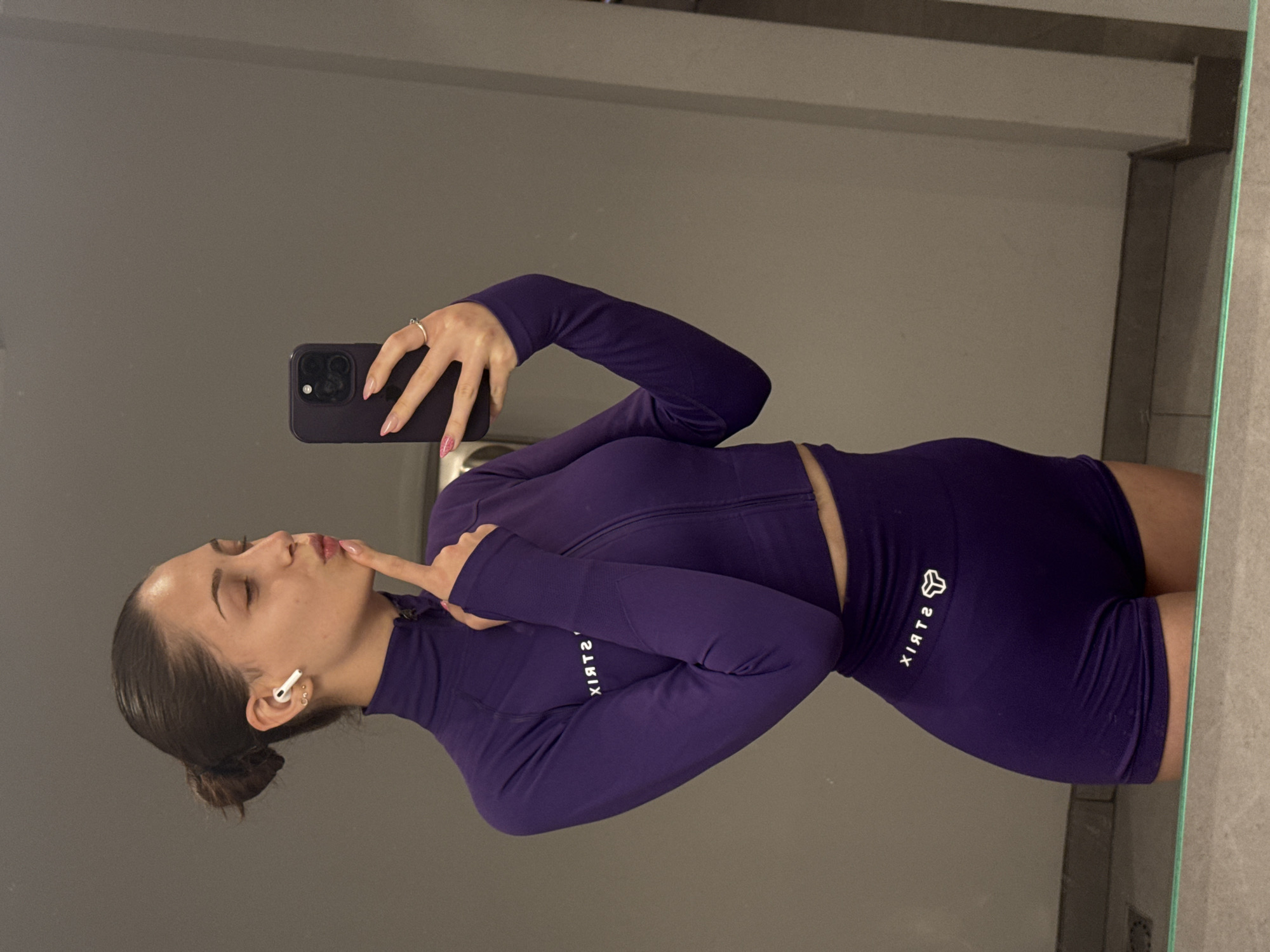 A young woman is taking a mirror selfie in a bathroom. She is wearing a purple athletic outfit consisting of a long-sleeved crop top with a zipper and matching shorts. The outfit has a white logo with geometric shapes and the text "STRIX" on the shorts. She has her hair pulled back in a bun and is wearing white wireless earbuds. Her eyes are closed, and she has her finger to her lips, giving a playful expression. A ring is visible on her right hand. The background is a gray wall, and a mirror reflects her image and the surroundings, revealing a tiled wall and a light fixture. The lighting suggests it is daytime or that the bathroom lights are on. The overall impression is casual and confident.
