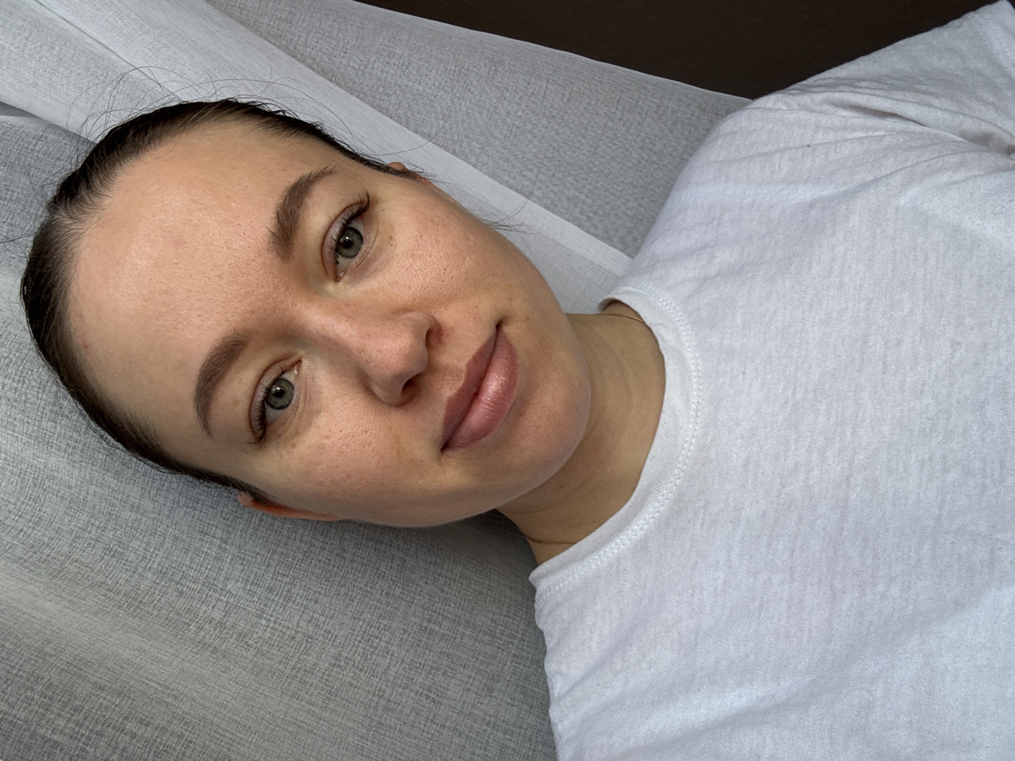 A woman lies at an angle against a light gray or white textured fabric, possibly a curtain or a sheet. She wears a white t-shirt. Her hair is pulled back. She has fair skin and light-colored eyes. The setting is indoors, with a dark brown or black surface visible in the background. The location is Ферма, Russia, but that information is not discernible from the image alone.