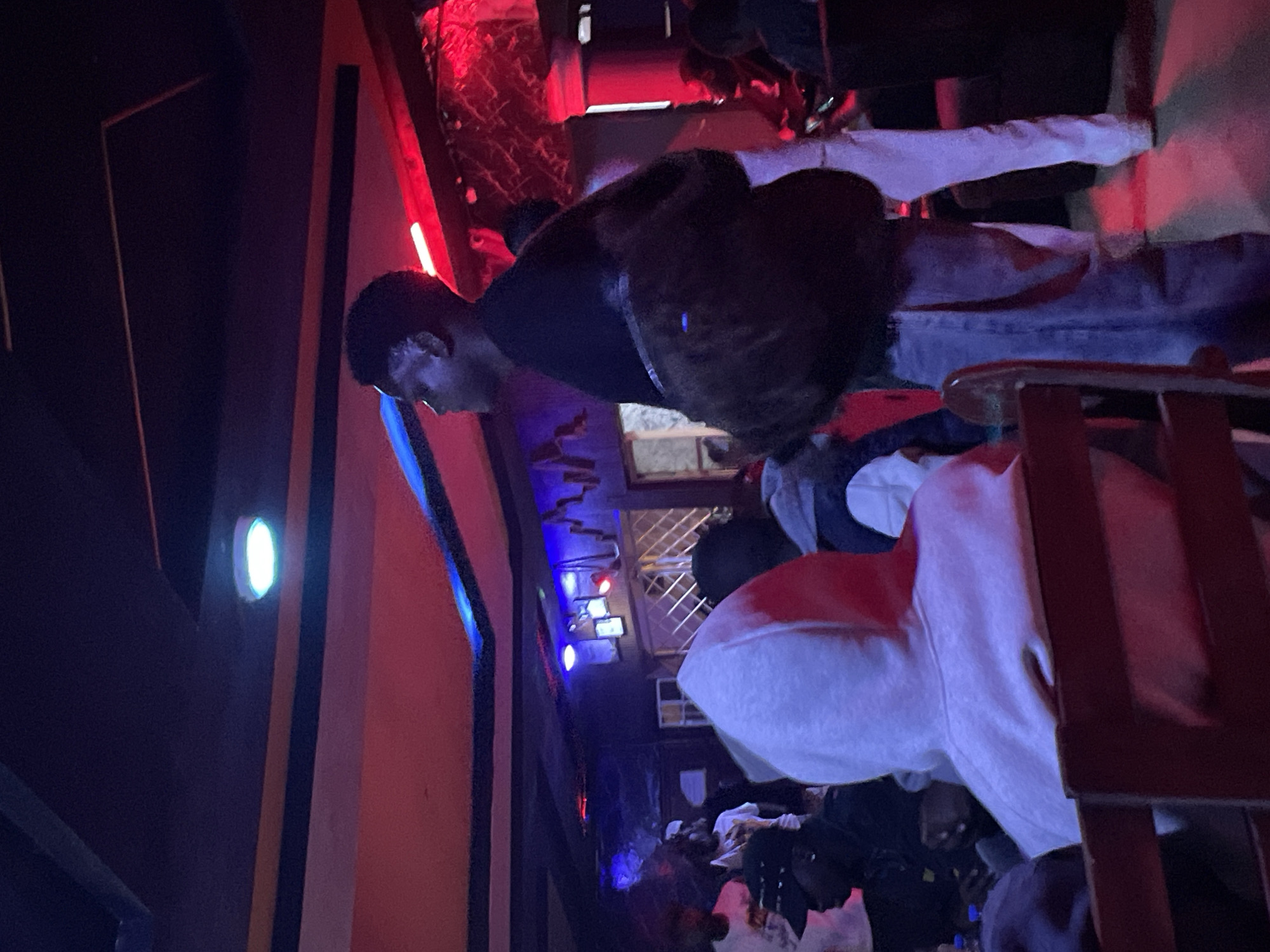 The image depicts an indoor scene with a group of people, possibly at a bar or club. The lighting is dim and predominantly red and blue, creating a vibrant, almost neon atmosphere. In the foreground, the back of a person wearing a light-colored hooded sweatshirt is visible, seated on a wooden chair. To the right, a young Black man is standing, facing away from the camera, wearing a black shirt and jeans, with a dark jacket draped over his shoulder. Other individuals are partially visible in the background, some standing and others seated. The environment appears to be a public space with a patterned ceiling and decorative elements on the walls. Based on the provided context, this scene is located in Durumi, Nigeria.