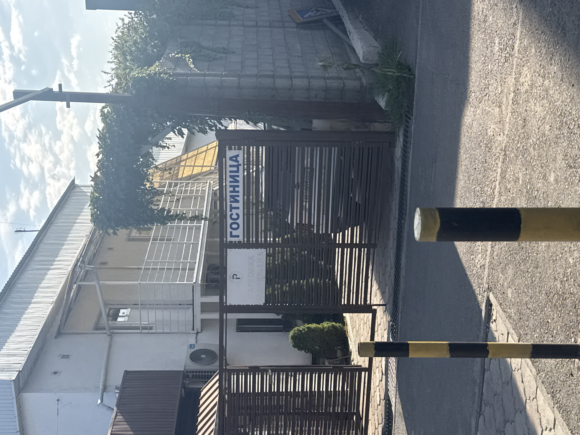 The image shows a building with a white facade and a corrugated metal roof. There is a dark metal fence in front of the building, with a gate that is partially open. Two signs are visible on the fence. One sign has a "P" symbol and the word "ПАРКОВКА" (PARKING) in Russian. Another sign has the word "ГОСТИНИЦА" (HOTEL) in Russian. There are two black and yellow striped bollards in the foreground. The environment appears to be a street or driveway leading to the hotel. The sky is visible in the background with some clouds. The scene is a street view of a hotel entrance in Bishkek, Kyrgyzstan.