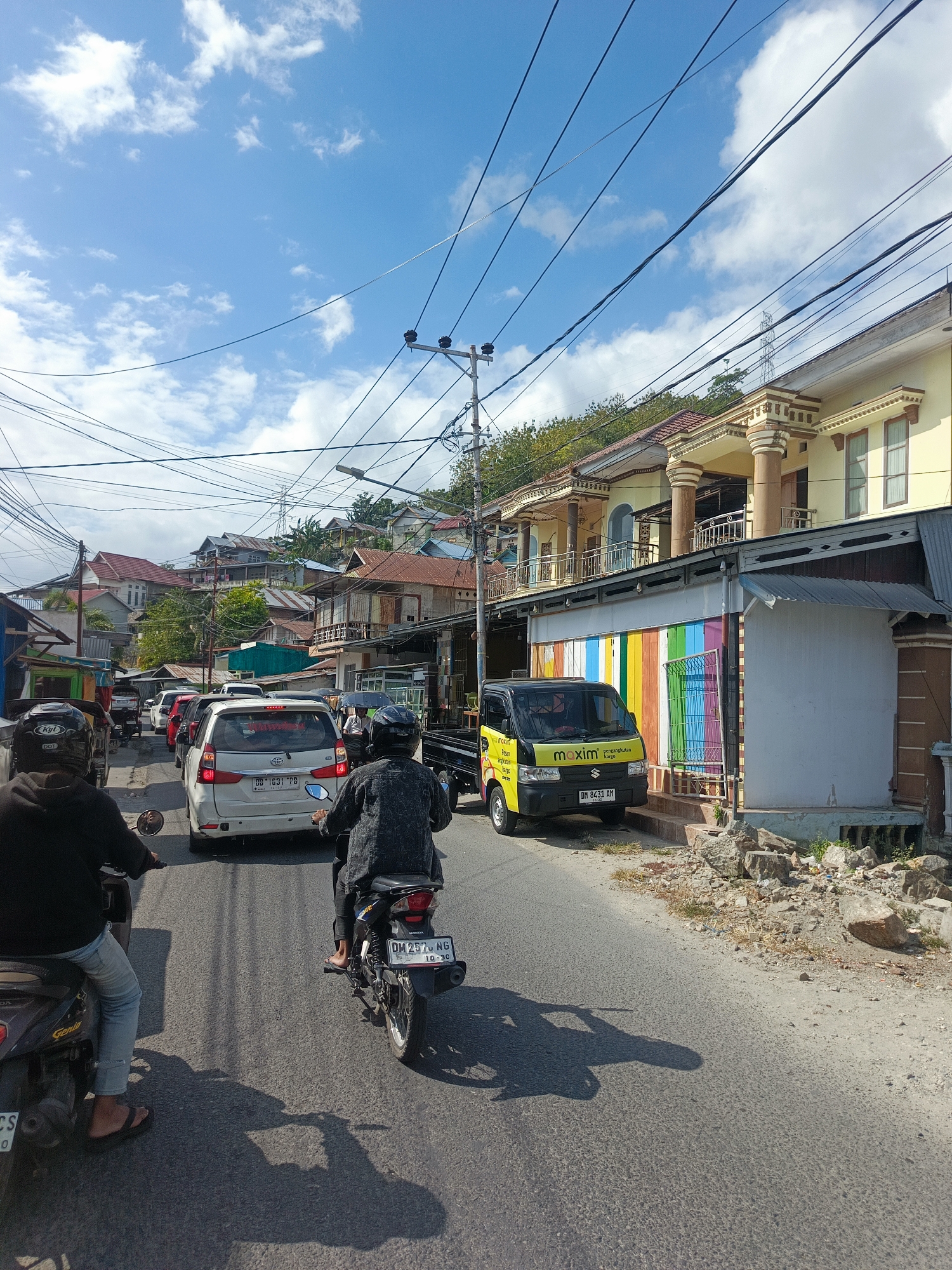 The image depicts a street scene in Gorontalo, Indonesia, characterized by moderate traffic and a mix of residential and commercial buildings. Two individuals are seen riding motorcycles in the foreground, one with a black jacket and the other in a dark grey patterned jacket. Behind them, traffic includes a white van with the license plate DB 1331 PB, and several other vehicles. A yellow Maxim delivery van with the license plate DM 8431 AM is parked on the right side of the road, in front of a building with a colorful, striped facade. The buildings in the background are varied, with some appearing to be houses and others more like small businesses, situated on a hillside. Overhead, a complex network of electrical wires crisscrosses the sky, indicating the urban setting. The sky is blue with scattered white clouds, suggesting a sunny day. The road surface is asphalt, with visible tire tracks and shadows cast by the vehicles and pedestrians. The environment appears to be an active, populated area within the city.