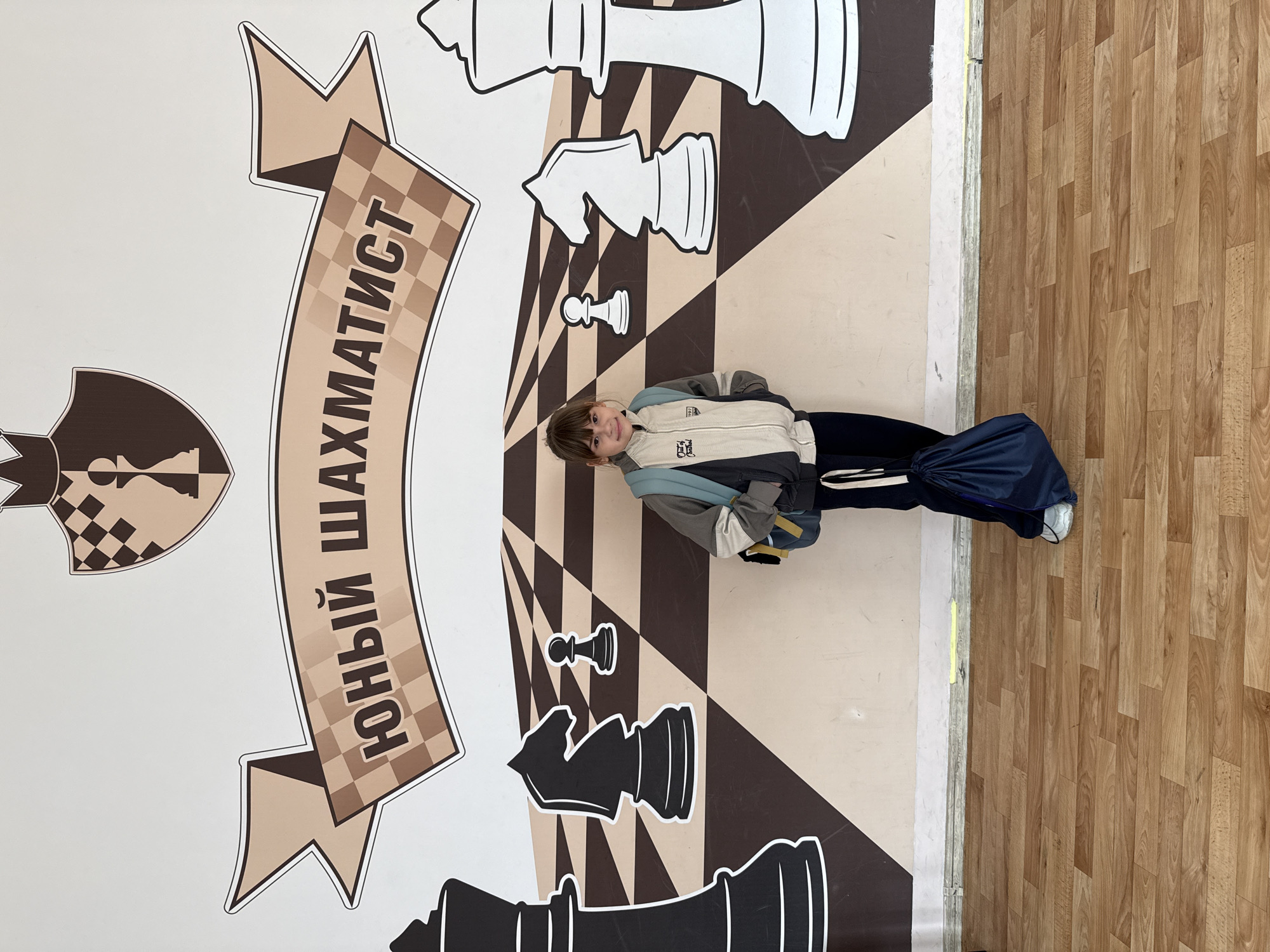 A young person stands on a floor with a large chess-themed graphic in what appears to be an indoor space. The graphic includes oversized chess pieces (king, knight, pawn, rook), and a banner in Russian reads "ЮНЫЙ ШАХМАТИСТ" (Young Chess Player). The surrounding floor is wood laminate. The specific location in Rusanovo, Russia, is not discernable from the image.