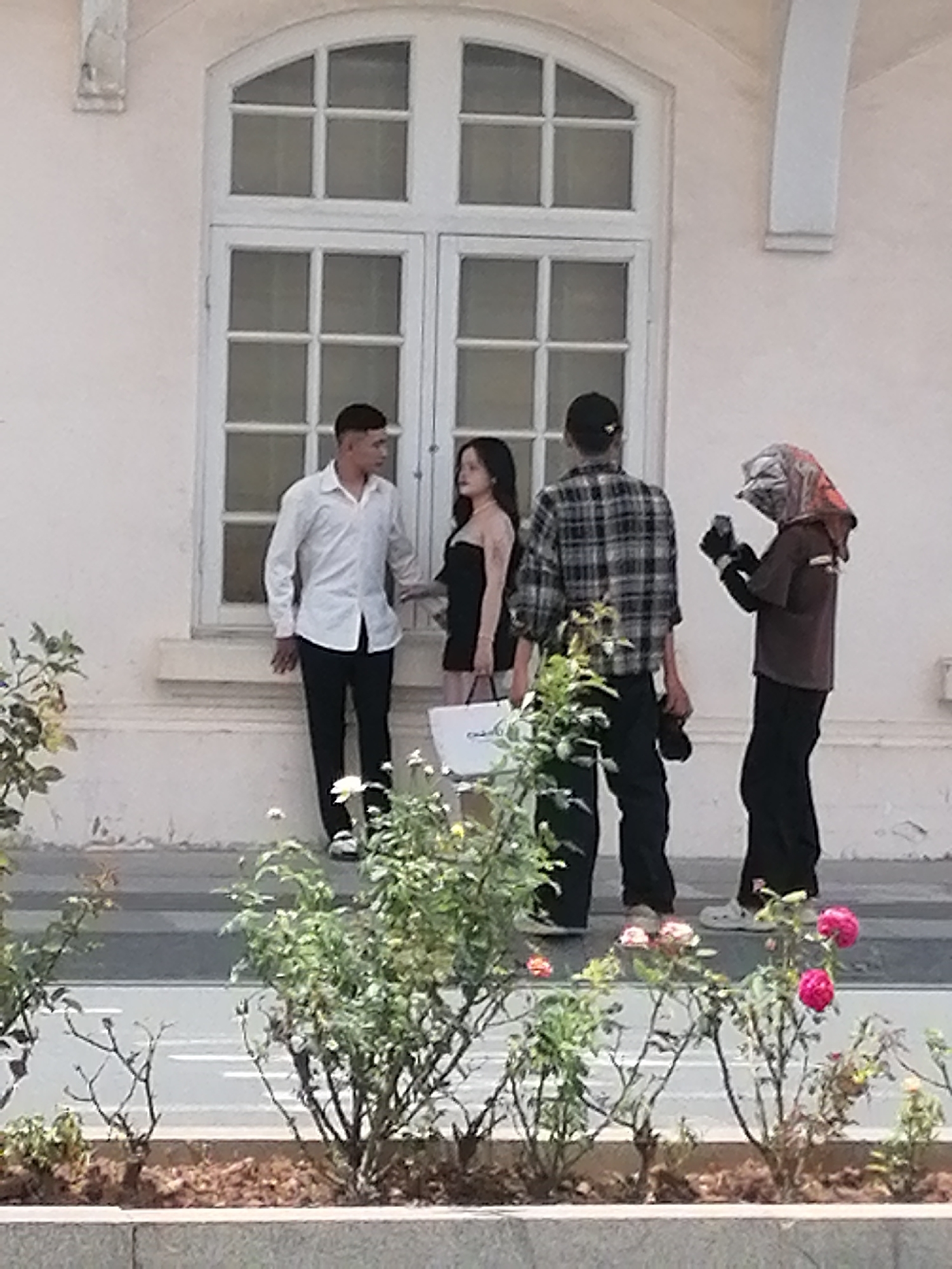In Da Lat, Vietnam, four individuals are positioned in front of a large, white, multi-paned window on a light-colored building. On the left, a man in a white long-sleeved shirt and black pants stands facing a woman in a black dress. The woman holds a white shopping bag. To the right of the couple, a person in a plaid shirt and black pants faces away from the camera, holding what appears to be a pair of shoes. Further to the right, another individual, partially obscured by a patterned head covering, holds a dark object, possibly a camera or phone, and is looking downwards. In the foreground, a variety of green foliage and pink roses are visible, suggesting a garden or landscaped area.