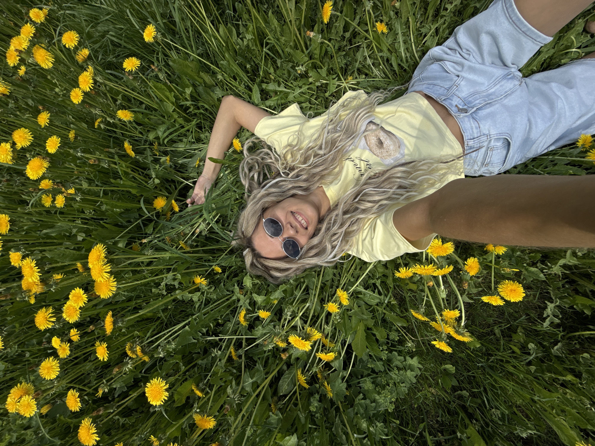 A woman lies on her back in a field of dandelions. She is wearing light blue jeans, a yellow T-shirt, and sunglasses. Her hair is long and blonde. She is smiling and looking up at the camera, which is presumably being held by someone above her. Her right arm is outstretched. The location is Unknown, Unknown.