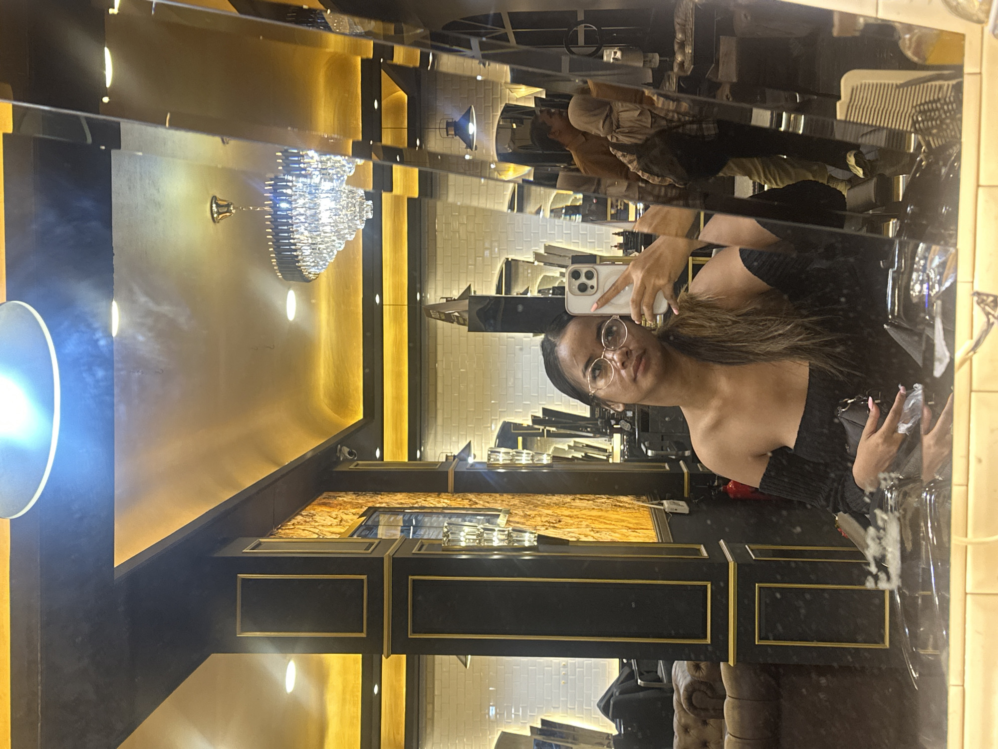 The image captures a mirror selfie taken by a woman in an upscale indoor setting. She is seen wearing glasses and a dark, off-shoulder top, with long, dark brown hair. She holds a light-colored smartphone with multiple cameras (likely an iPhone) to take the picture, with a neutral expression looking towards the phone.
The background, reflected in the mirror, reveals a luxurious interior. Dominant decorative features include dark, glossy wall panels framed with gold accents. Some panels display a light brown, marble-like pattern, while others appear as dark, plain surfaces. A prominent, ornate crystal chandelier hangs from the ceiling, contributing to the sophisticated ambiance. Additional ambient lighting comes from spotlights and a circular wall sconce, illuminating the golden-toned walls and ceiling.
In the deeper reflection, other individuals, likely patrons or staff, are visible, along with sections of white subway tile-patterned walls and cushioned seating, possibly a sofa or chair. The overall atmosphere is elegant and warm, suggesting a high-end venue such as a salon, hotel lobby, or restaurant. The setting is indoors, with artificial lighting, so the time of day is not explicitly indicated but appears to be evening or late afternoon. No discernible text is visible within the image. Given the provided context, the location is North Jakarta, Indonesia.