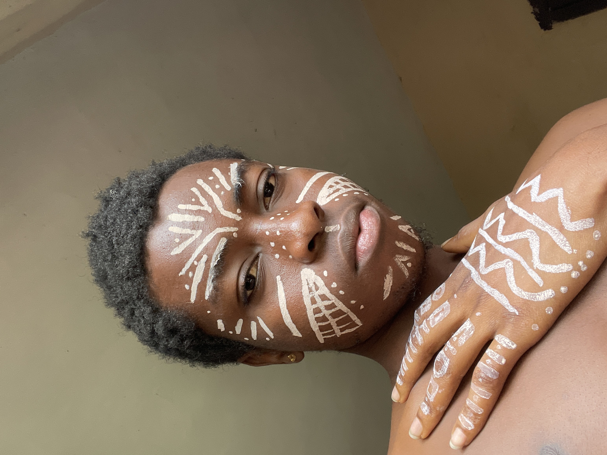 A medium close-up, portrait-style shot captures a young Black man with intricate white body paint adorning his face and right hand. He looks directly at the viewer with a calm and serious expression, his lips slightly parted.
The white paint forms various geometric and abstract patterns. On his forehead, there are diagonal lines, dots, and zig-zag motifs. Around his eyes and nose, more lines and dots are visible, with a distinctive triangular shape featuring an internal grid pattern on his right cheek (viewer's left). His chin and jawline also feature scattered dots and lines. His hair is short, dark, and curly. A small, gold stud earring is visible in his left ear.
His right hand is positioned across his left shoulder or upper arm, also decorated with prominent white zig-zag lines and smaller dots on the fingers and across the back of the hand. The skin tone of the subject is dark brown.
The background is a plain, light brown or beige wall, suggesting an indoor setting. The lighting appears soft and even, indicating daytime but without direct harsh sunlight. There is no visible text in the image. The image was taken in Nkpor, Nigeria.