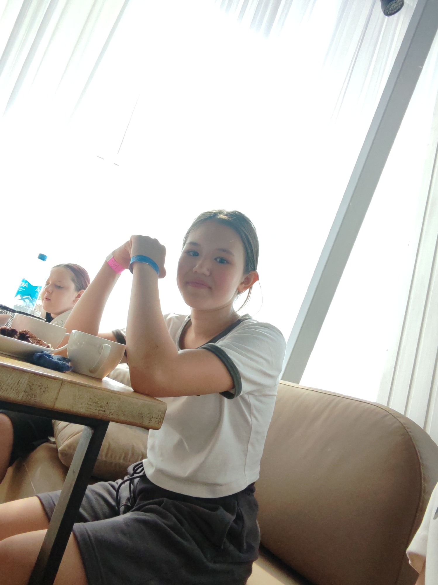 A young East Asian girl, with dark hair pulled back, sits at a wooden table, looking at the camera. She is wearing a white t-shirt with a dark trim around the collar and sleeves, and dark gray shorts. On her left wrist, she has a pink wristband, and on her right wrist, a blue wristband. Her arms are crossed in front of her, with her hands clasped. To her left, a white mug and a plate with what appears to be dark food are on the table. Behind her, a younger girl with reddish-brown hair is partially visible, looking to her left. A bottle of water is also on the table. The background is dominated by large, bright windows with sheer white curtains, and a light-colored wall or partition. The setting appears to be indoors, possibly a cafe or dining area, with a couch visible on the right side of the image. The lighting is bright, coming from the windows. The image does not provide any specific information to indicate a location in the city of Amursk, Russia.
