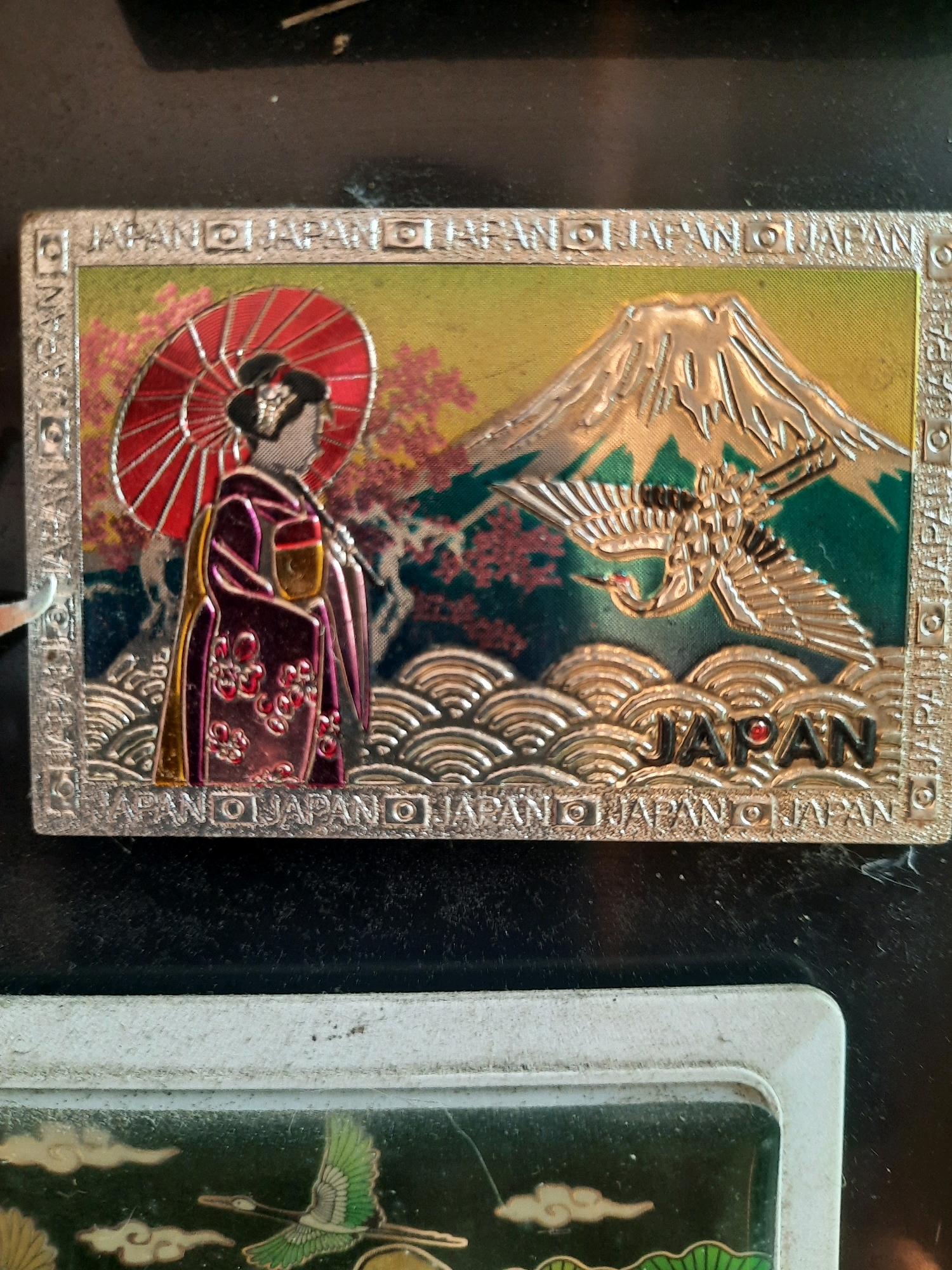 The image shows a close-up of a rectangular magnet with a Japanese theme. The magnet features a scenic depiction of Mount Fuji with a crane flying in front of it, and a woman in a kimono holding a red umbrella. The word "JAPAN" is prominently displayed in black letters with a red dot accent. The border of the magnet is adorned with the word "JAPAN" repeated several times, interspersed with decorative elements. The magnet appears to be attached to a dark, textured surface, possibly a refrigerator or a metal cabinet. The scene is well-lit, with the magnet being the primary focus. The background is dark and out of focus, suggesting that the photograph was taken in an indoor environment. There are no people visible in the image. The overall impression is that of a souvenir item. There is no indication of the location being in East Jakarta, Indonesia.