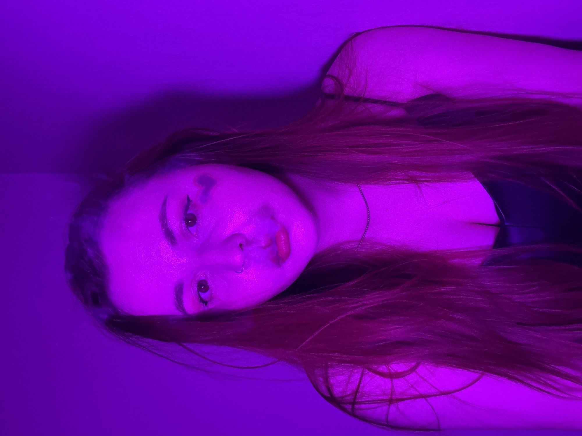 A female person with long, reddish-brown hair and fair skin is lying down. She has dark eyeliner and a nose piercing. She is wearing a black top with thin straps. The scene is illuminated with a strong purple light, obscuring details. No information is available to confirm the location in Izhevsk, Russia.