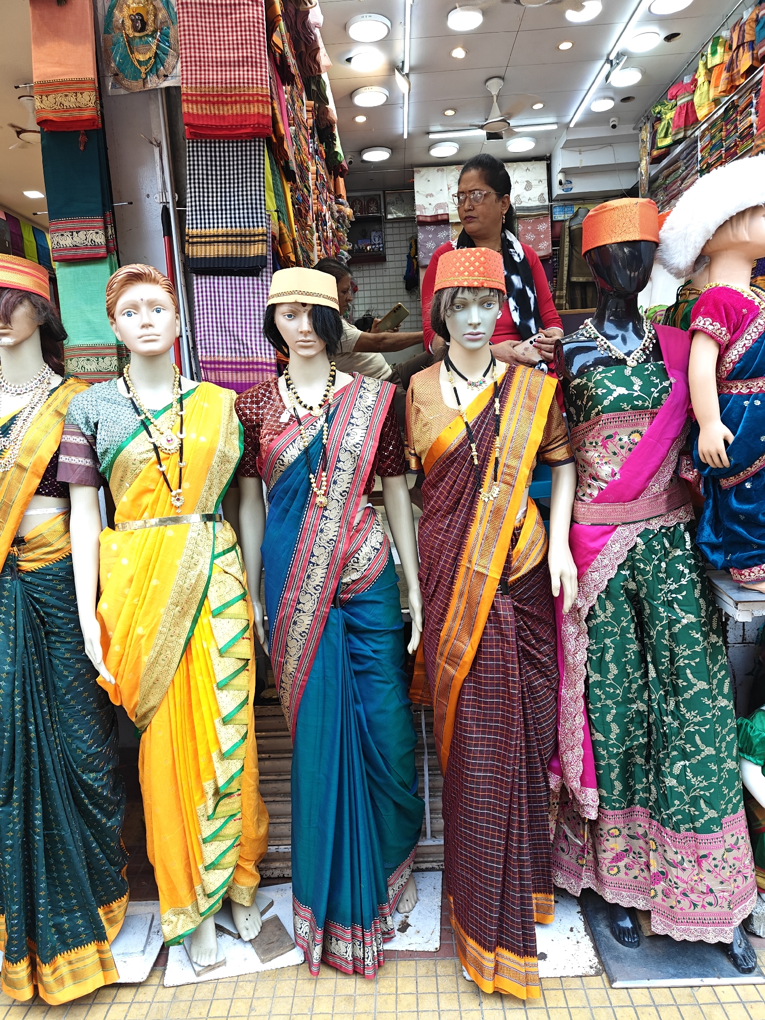 The image depicts the storefront of a textile shop, located in the city of Unknown, Unknown. Five mannequins are positioned at the entrance, displaying traditional Indian garments.
From left to right:
1. A partially visible mannequin wears a dark teal saree with gold borders and small motifs, accessorized with a gold-colored necklace and a reddish-orange cap.
2. A full-body mannequin with light brown hair and blue eyes wears a bright yellow saree with green and gold borders, draped over a light green blouse. It is adorned with a gold-colored waist belt and a long black and gold beaded necklace.
3. A full-body mannequin with dark hair wears a dark teal saree with red, black, and gold patterned borders, draped over a brown blouse. It is accessorized with a long black and gold beaded necklace and a light beige cap.
4. An upper-torso mannequin wears a dark reddish-brown and orange plaid-patterned saree with intricate gold borders, draped over a brown blouse. It is adorned with a long black and gold beaded necklace and an orange cap.
5. A partially visible child mannequin wears a green lehenga (skirt) with intricate gold, pink, and red floral embroidery, paired with a matching pink dupatta and a dark teal blouse. It is accessorized with a white pearl-like necklace and a white fluffy hat.
Inside the shop, positioned behind the central mannequins, a female individual with dark hair, glasses, and a red top stands, observing towards the right side of the frame. The shop interior showcases numerous textiles, including stacks of colorful sarees and vertically hung checkered fabrics in red/white, black/white, and purple/white patterns. A golden deity figure is mounted high on the left wall, and a security camera is visible on the upper right. The ceiling features multiple recessed lights and a white ceiling fan. The mannequins stand on white or dark platforms on a yellow-brown tiled floor at the shop's threshold.