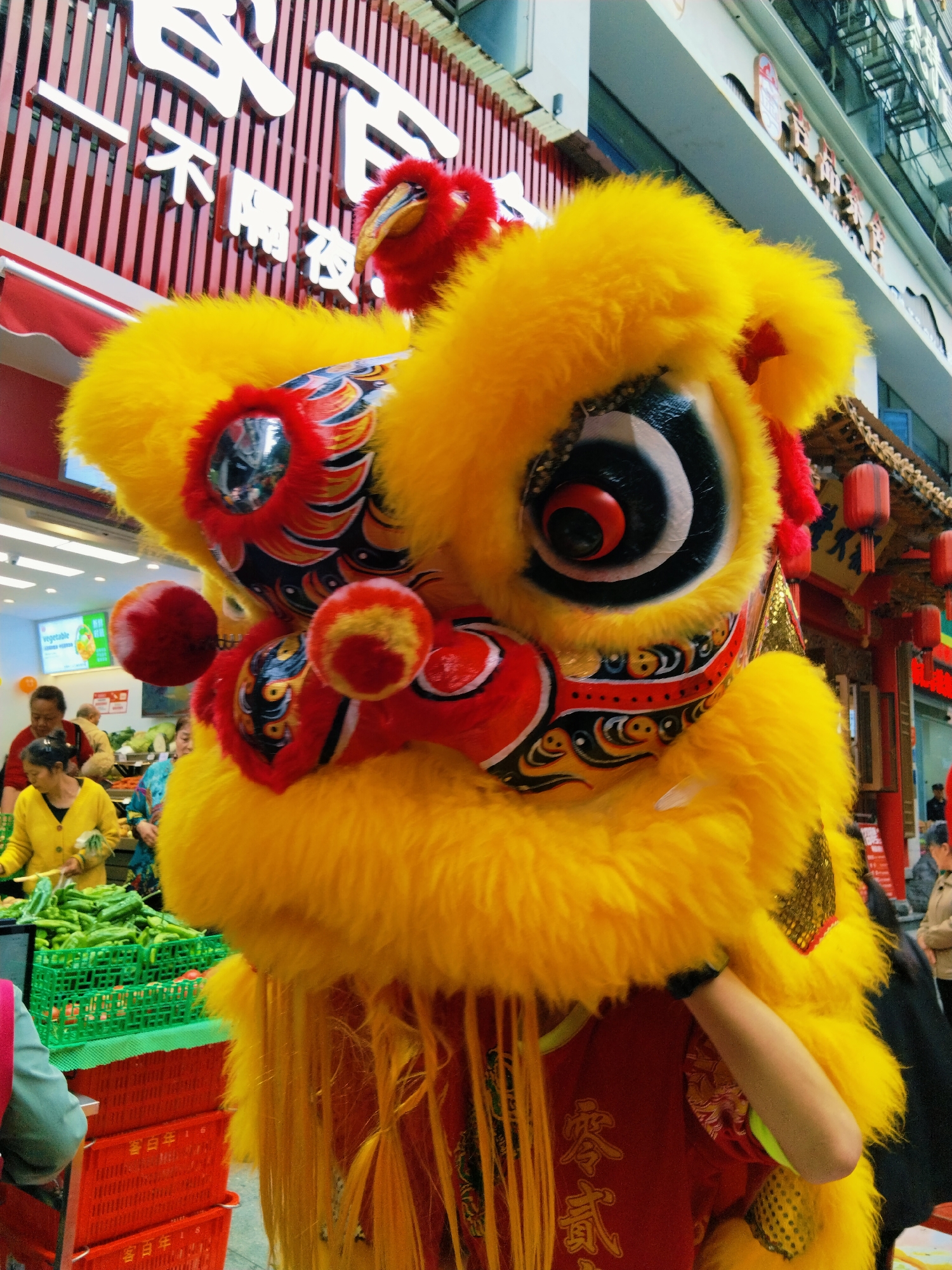The image depicts a vibrant yellow lion dance costume, a traditional performance art form often associated with Chinese New Year and other celebrations. The costume is elaborate, featuring a large, furry yellow head adorned with intricate patterns in red, black, and gold. A person's arm is visible within the costume, suggesting an ongoing performance.
The background reveals a bustling urban environment. In the left side of the frame, people are browsing produce in a market setting. The presence of fruits and vegetables and the bright store lights suggest a marketplace or a grocery store. Red crates are stacked, indicating the movement of goods. The architecture in the background, with red lanterns and Chinese characters, indicates a location within Yuzhong District, China, where traditional architecture blends with modern city life.