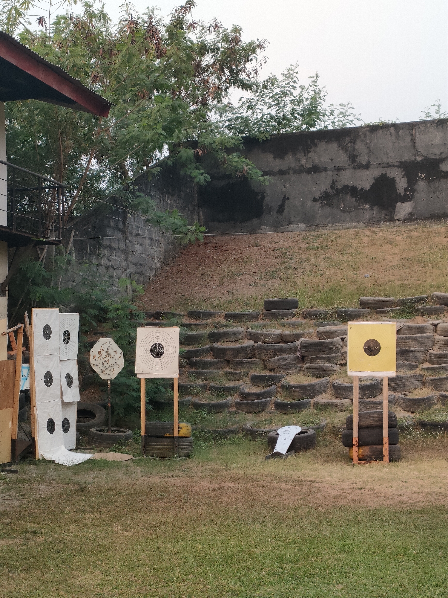 The image shows an outdoor shooting range with multiple targets set up on a grassy area. In the background, a large concrete wall forms a berm, with stacked tires serving as additional barriers and seating or a visual divider. The overall impression is of a functional, if somewhat rustic, shooting facility. There are no people visible in the image.