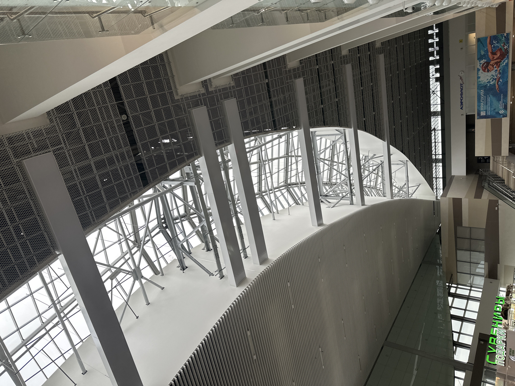 The image shows the interior of a modern, large building with a high, multi-level ceiling featuring a combination of glass, metal, and paneled surfaces. The ceiling structure is complex, with visible metal trusses and beams supporting a large skylight that allows natural light to enter. Below the skylight, there are sections of dark, perforated metal panels and white, curved, or angled architectural elements.
In the lower part of the image, the building's interior architecture continues with large glass windows and sections of ribbed paneling. There are also signs visible, including one with Russian text that reads "ПОДАРКИ" (Gifts) in green lettering and another larger banner featuring the "AEROFLOT" logo and "УРАЛХИМ" (URALCHEM) text, along with an image of a swimmer. The environment appears to be a public indoor space, possibly an airport or a large commercial complex. The lighting is a mix of natural light from the skylight and artificial light sources that are not clearly visible. The scene is clean and appears to be unoccupied by any people in the frame, with the exception of a distant, small figure visible on the stairs in the lower right quadrant. The overall impression is one of a spacious and architecturally striking interior. The provided context indicates this location is in Bolshoye Savino, Russia.