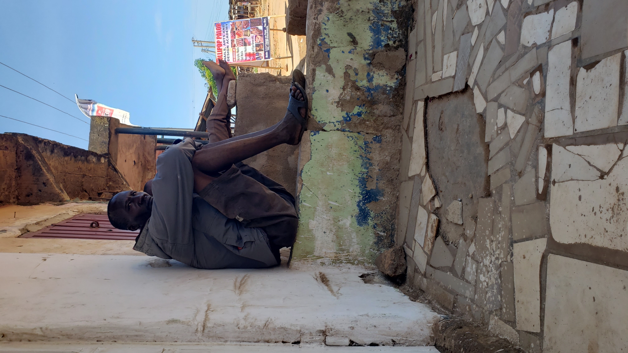 The image depicts an outdoor scene in Sabon Tasha, Nigeria, under a clear blue sky with ample daylight. An adult male of dark complexion is seated on a low concrete ledge adjacent to a building on the left. He wears a light grey hooded jacket and dark, soiled shorts. His knees are bent, with his arms wrapped around them. His gaze is directed forward, slightly towards the viewer. His right foot, clad in a dark sandal, rests on the ledge, while his left bare foot rests on a small, light-colored stone positioned on the ledge. The building wall behind him is white, exhibiting significant dirt, wear, and patches of discoloration. Directly behind the man, a reddish-brown, vertically slatted shutter is visible. The concrete ledge he occupies features peeling green and blue paint over a rough, unpainted base. Above the ledge, a rough, unpainted concrete overhang extends from the building. The ground in the foreground consists of irregularly shaped, light-colored tiles, many of which are broken, cracked, or missing, revealing underlying dirt. In the mid-ground to the right, a white A-frame sign is prominent, displaying "LOLLIPOP PALOR" and "NAILS, ARTS AND DESIGN ACADEMY." Listed services include "Acrylic Nails," "Gel Nails," "Normal Nails," "Pedicure," and "Carving of Eye Brow." The motto "Motto: S.O.P Swag on Point - You Man" is visible at the bottom of the sign. Power lines are visible stretching across the upper right portion of the sky, with distant vegetation and additional structures in the background.