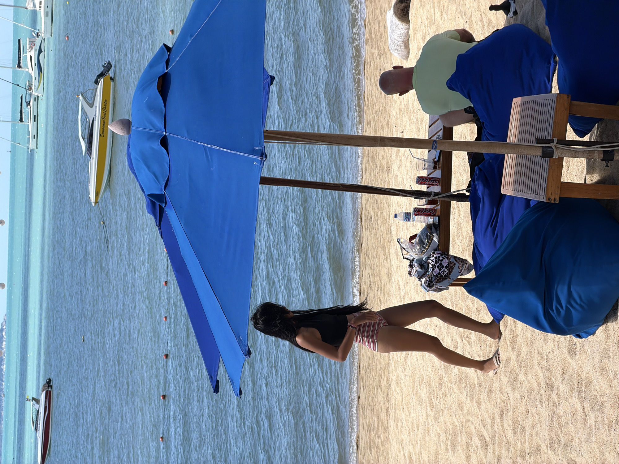 A woman with long dark hair, wearing a black tank top and striped shorts, stands on a sandy beach in Baan Bo Phut, Thailand. She is facing left, with her left hand near her hip. To her right, a man with a shaved head, wearing a light green t-shirt, sits on a blue beanbag chair, facing away from the camera. They are under a large blue umbrella, with a wooden table between them. The table holds several bottles of Coca-Cola, glasses, and a bag of items. In the background, the turquoise ocean is visible, with several boats and sailboats on the water. The scene appears to be a relaxed beach setting, likely during the day.