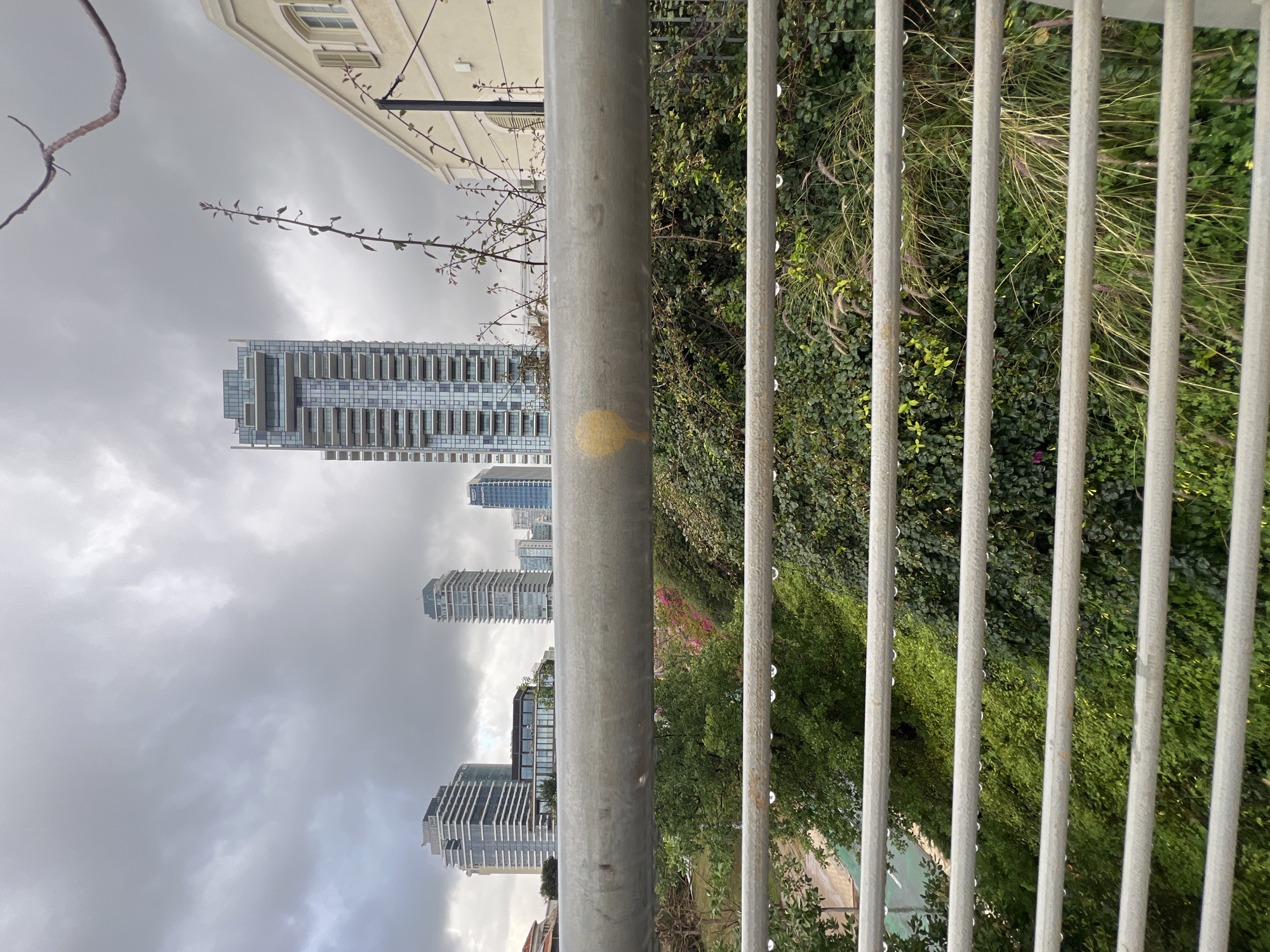 View from a pedestrian bridge in Tel Aviv, Israel. The perspective is from behind a silver metal fence and railing. Tall buildings are visible in the background against a cloudy sky. Green vegetation lines the area behind the fence. No people are apparent.
