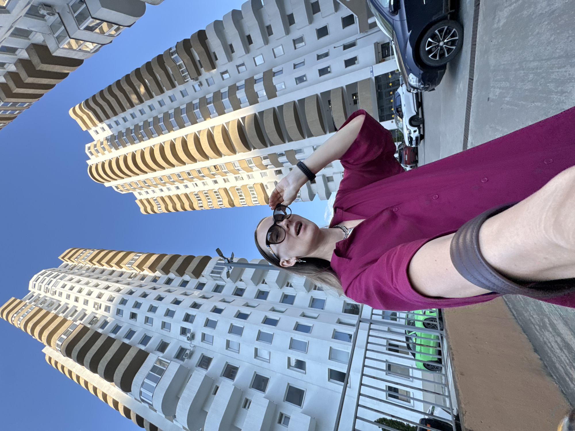 A selfie is taken by a woman wearing a magenta shirt and sunglasses, standing in front of two modern high-rise apartment buildings. The buildings, with their curved balconies and white facades, are characteristic of contemporary architecture in Sochi, Russia. A green car is visible in the background parked on a lower level. The sky is clear and blue, indicating a sunny day. The perspective of the photo is low-angle, emphasizing the height of the buildings.