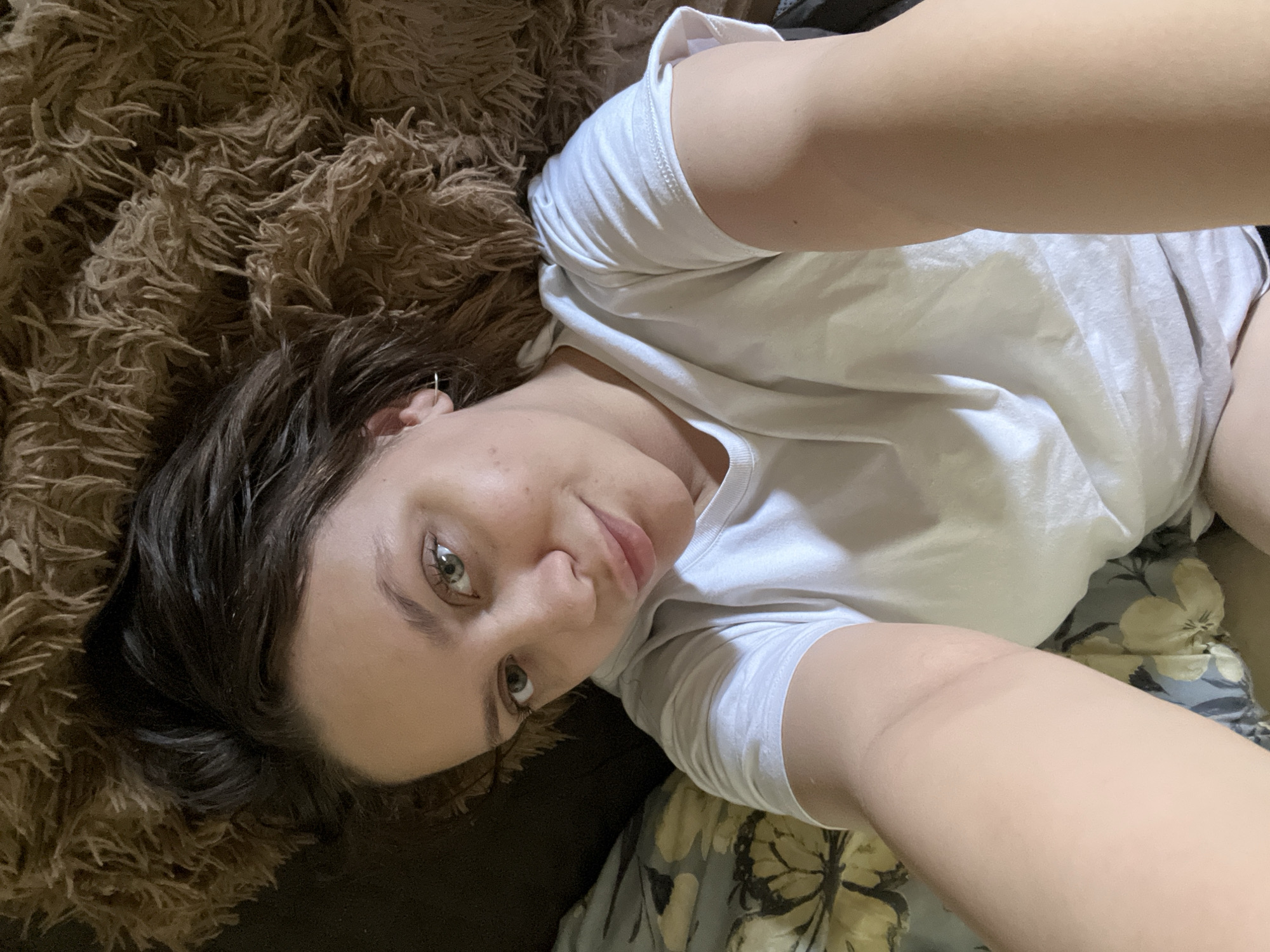 A young Caucasian woman, appearing to be in her late teens or early twenties, is captured in a close-up shot, likely a selfie. She has dark brown hair and captivating light green or hazel eyes, looking directly at the camera with a relaxed and slightly pensive expression. A small silver hoop earring is visible in her left ear.
She is dressed casually in a plain white short-sleeved t-shirt and light-colored shorts or underwear, suggesting a comfortable, at-home setting. The woman is lying down, with her head tilted, on what appears to be a bed or sofa. To her left, a plush, shaggy dark brown blanket or throw provides a textured backdrop. To her right, a light-colored fabric with a subtle floral or butterfly pattern is visible, possibly a pillowcase or another blanket. The lighting is soft and even, typical of an indoor environment during daylight hours. The overall mood of the image is intimate and casual, portraying a moment of relaxation. No text is visible in the image. This scene takes place in Chaikovsky, Russia.