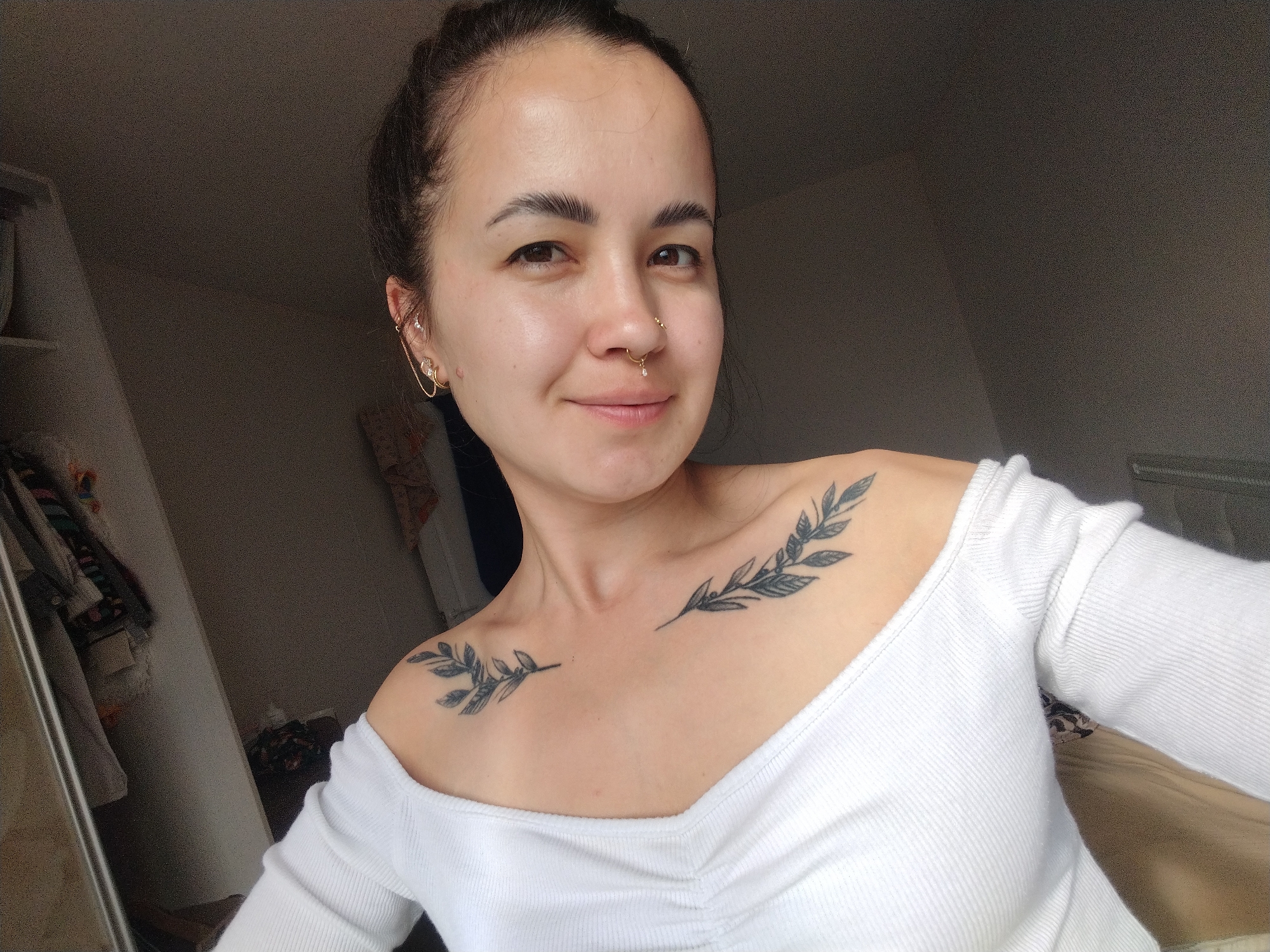A young woman with dark hair pulled back in a bun is shown in a close-up, upper-body selfie. She is wearing a white, ribbed, off-the-shoulder top and has a nose ring and earrings. She has two tattoos on her chest and collarbone area, depicting branches with leaves. The background is dimly lit and appears to be an indoor setting, possibly a bedroom, with a closet visible to the left. There is no indication of the city of Noviy, Russia, in the image.