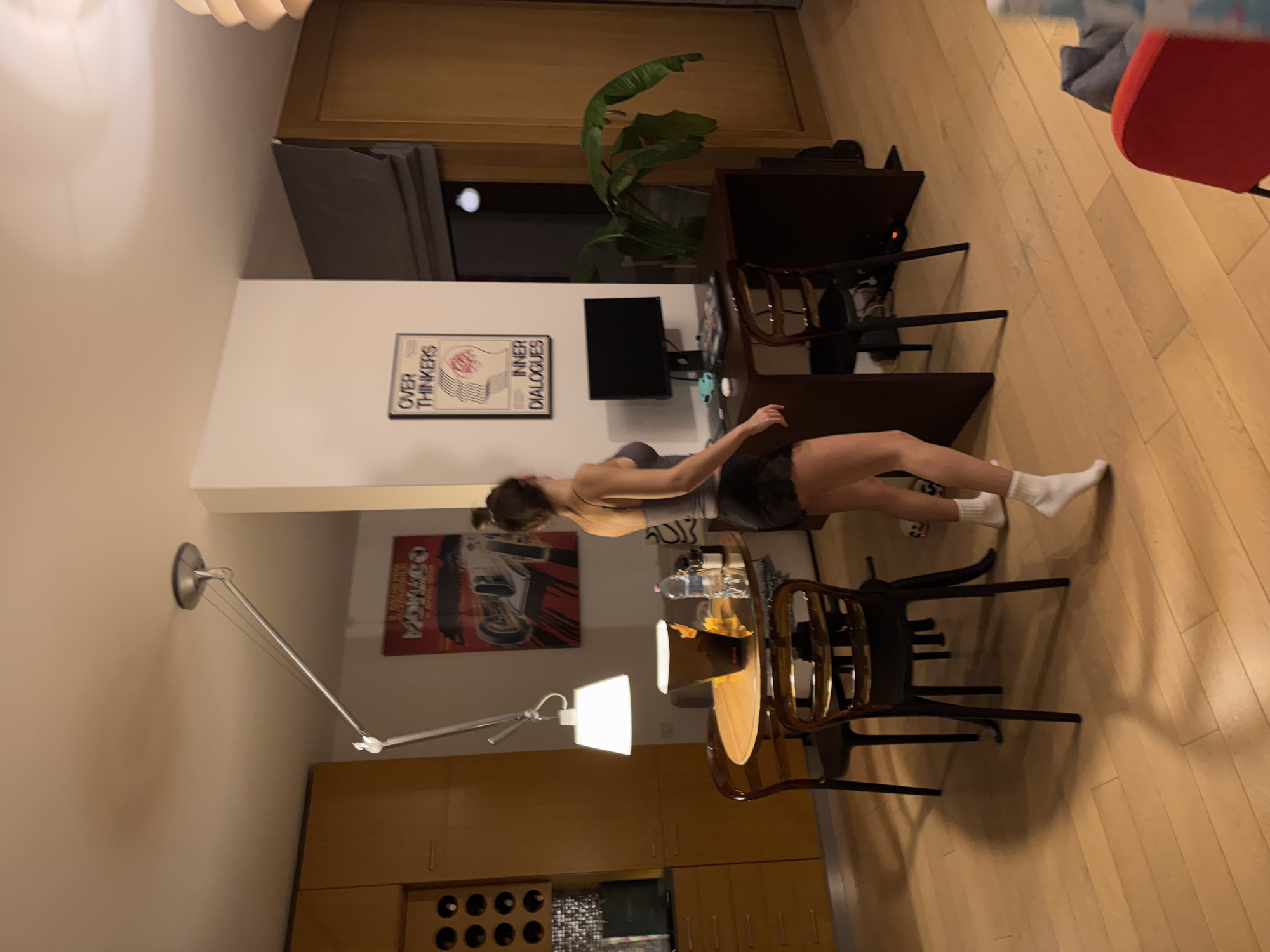 A woman is captured in a domestic interior, possibly in Warsaw, Poland. The location is identifiable by Polish poster art on the walls. She is standing and leaning over a dining table, which is surrounded by wooden chairs. A floor lamp illuminates the scene. The environment includes a TV, a plant, and various furniture pieces, giving the impression of a living and dining area.